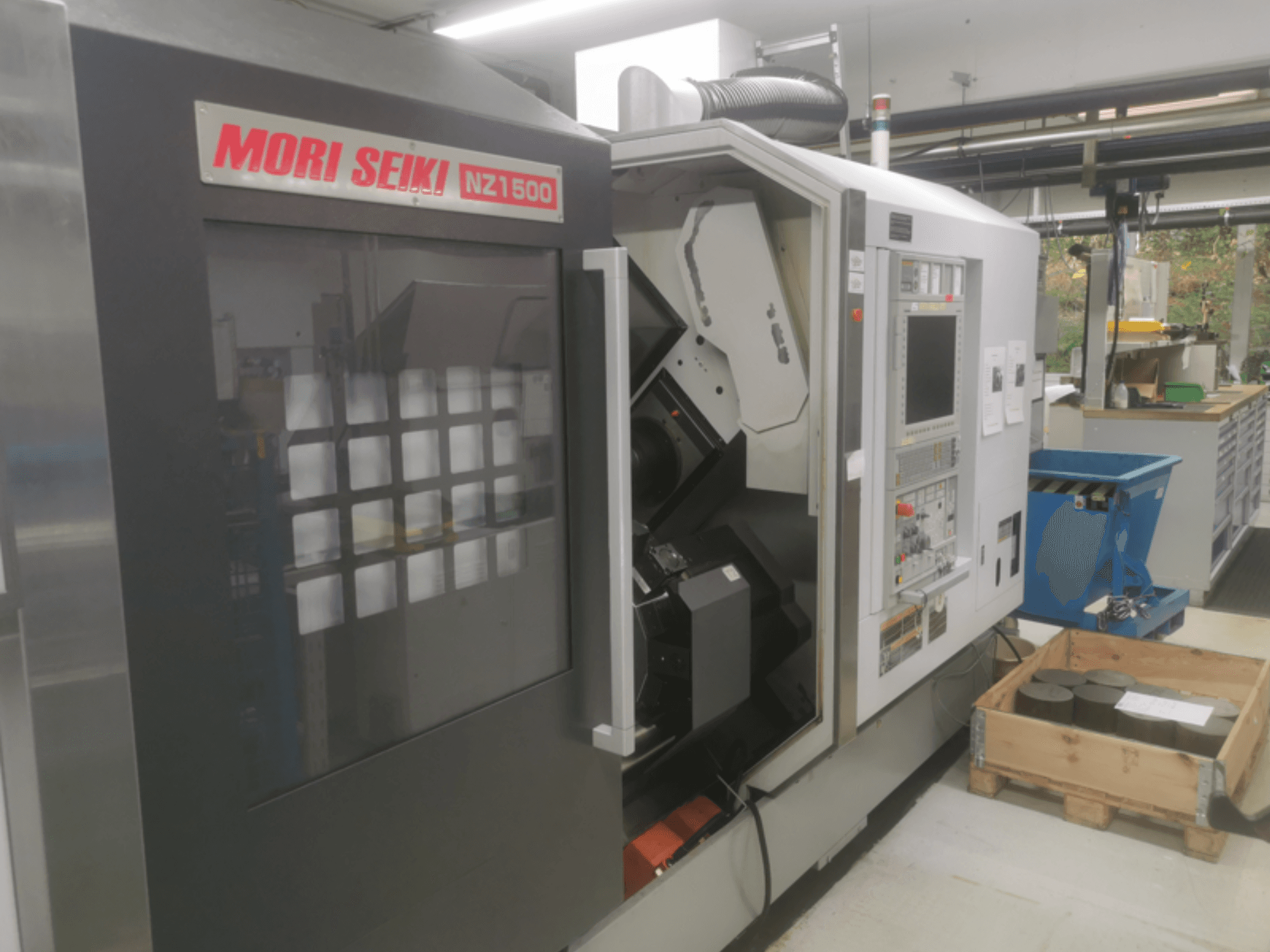 MORI SEIKI NZ1500 CNC mašīna nelielā leņķī, redzams vadības panelis un darba vieta ar metāla sagatavēm uz koka paliktņa.