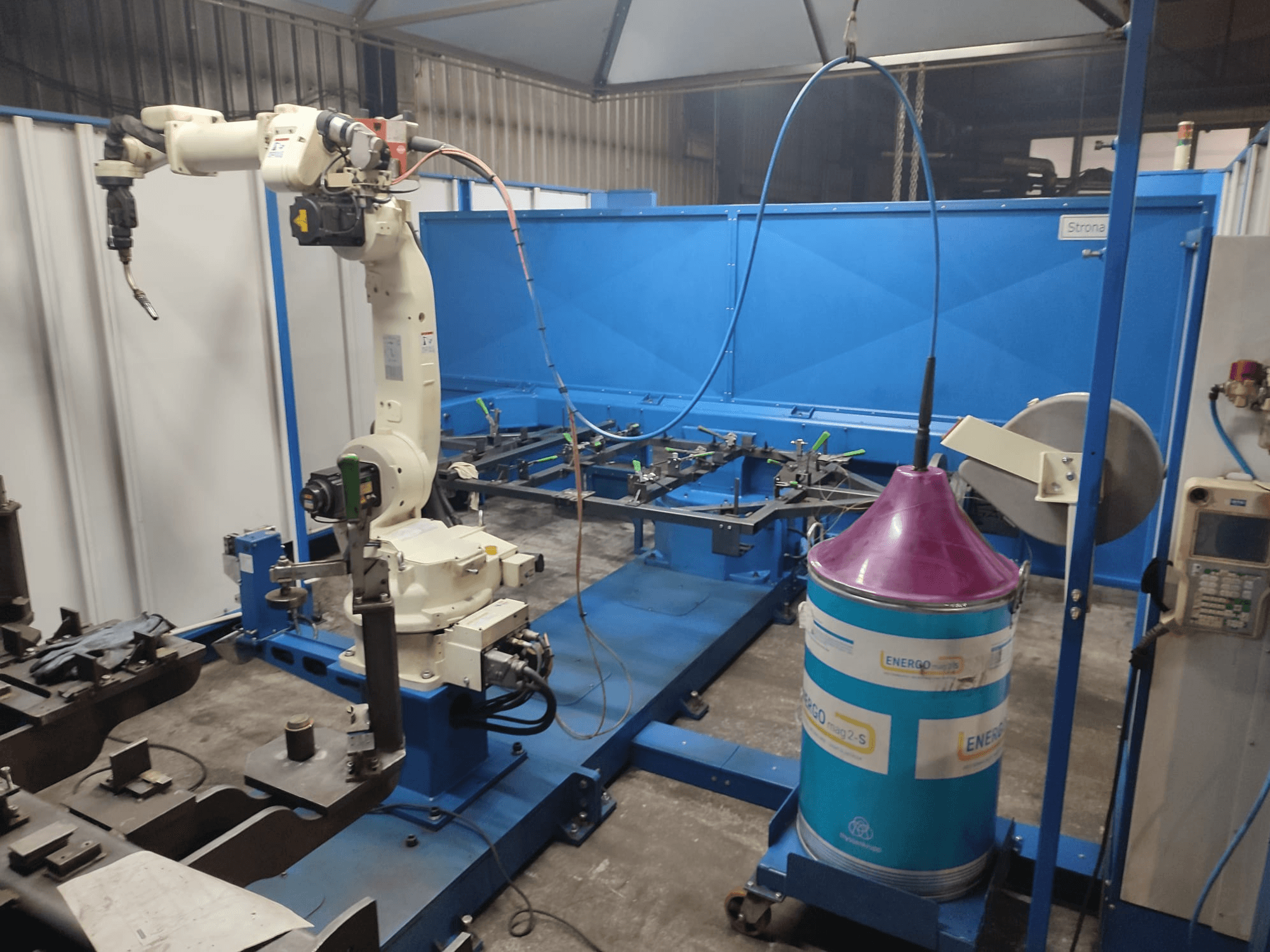 Rūpnieciska robotizēta roka ar baltu un zilu pamatni, uzstādīta darbnīcā ar zilu darba virsmu un metāla komponentiem.