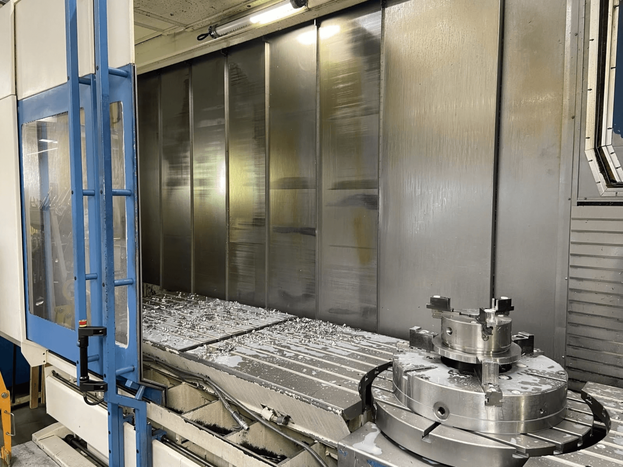 Vertikālais apstrādes centrs AXA VHC-3-3000-M/E, skats no priekšpuses, kurā redzamas nofrēzētās metāla skaidas uz darba galda un griešanas instrumenti.