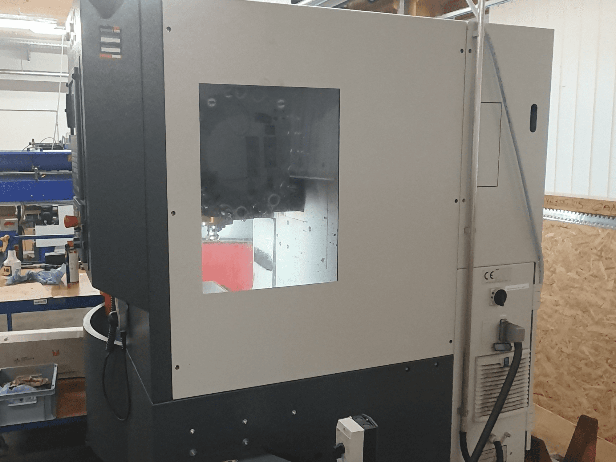 Metālapstrādes mašīnas sānu skats ar redzamu vadības paneli, lielu skatu logu un sarežģītām iekšējām detaļām.