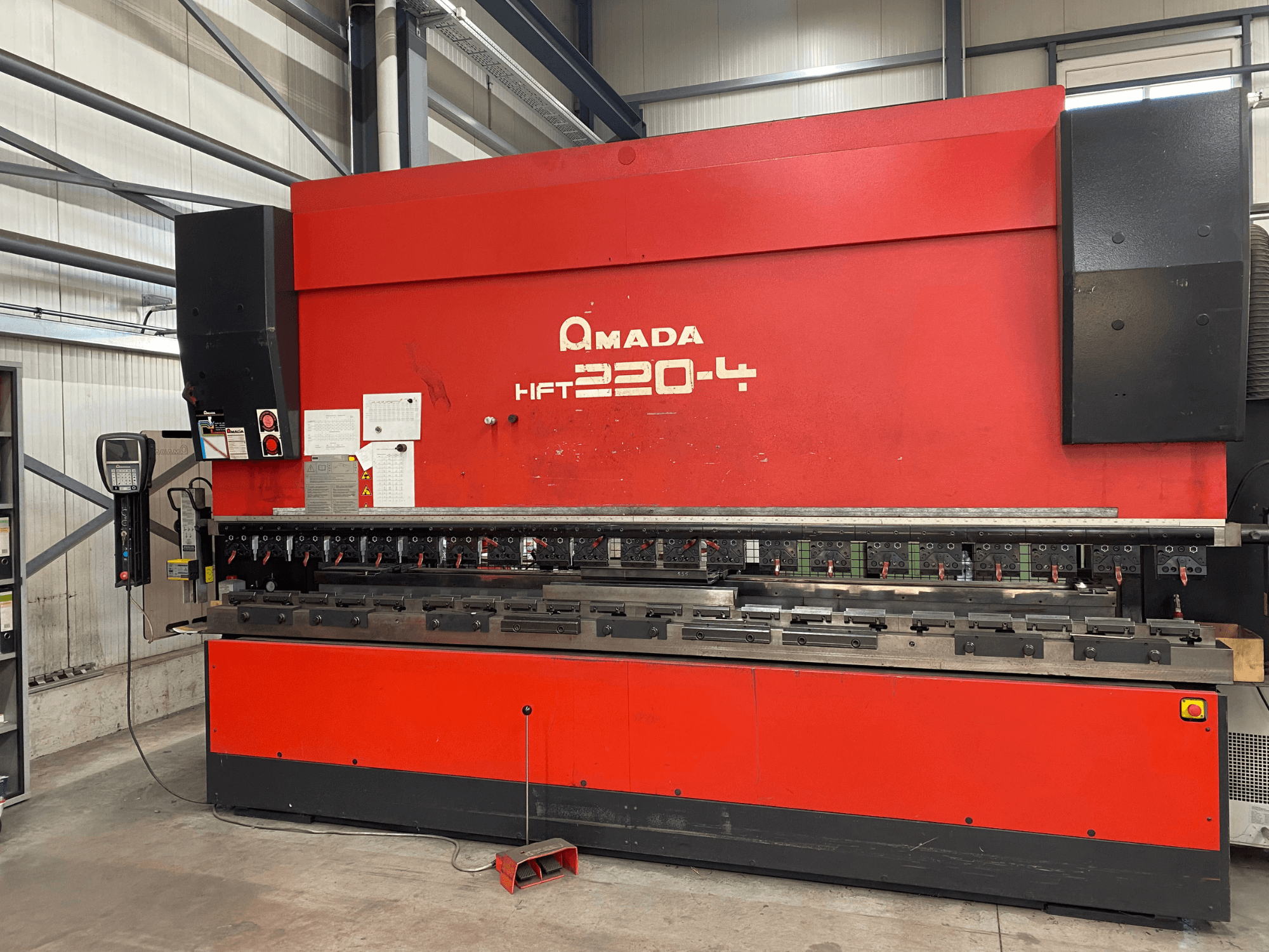 Sarkanā AMADA HFT 220-4 preses bremze rūpnīcā, priekšējais skats, kurā redzams vadības panelis un lieces instrumenti uz darba virsmas.