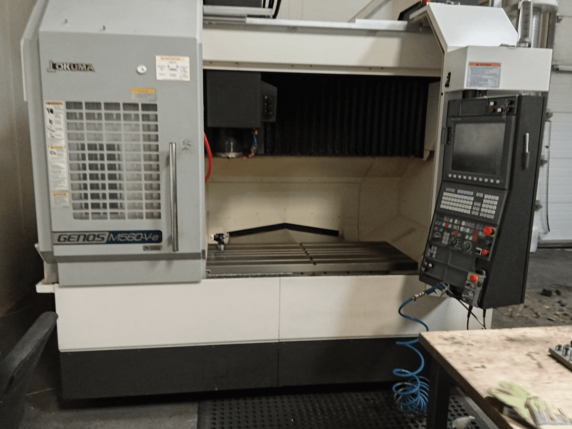 Okuma GENOS M560-V, vertikālā apstrādes centra priekšējais skats ar vadības paneli, CNC saskarni un redzamo griešanas zonu.