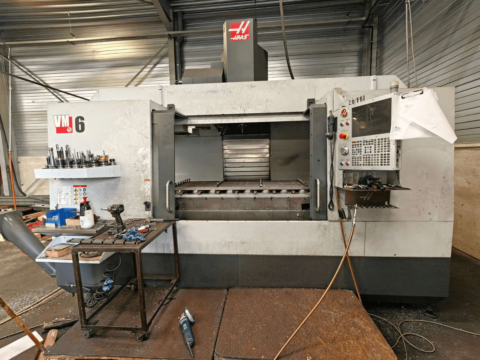 Vertikālais apstrādes centrs „Haas VM-6“, skats no priekšpuses, kurā redzama instrumentu glabātava, vadības panelis un darba zonā esošie atkritumi.