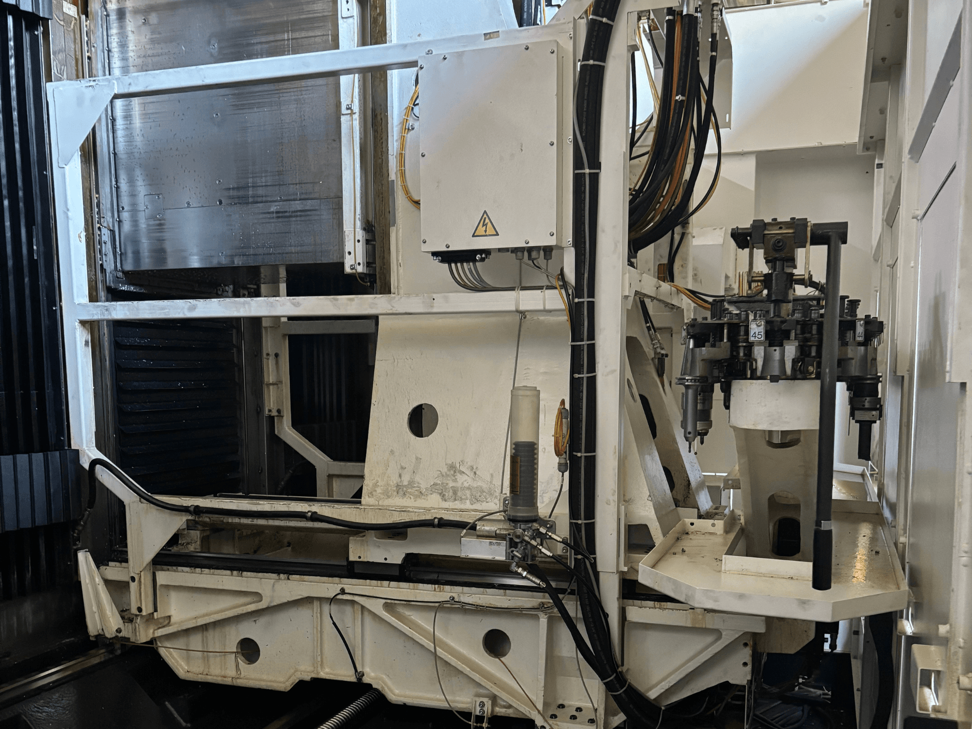 Rūpniecisko mašīnu sānu skats, kurā redzams balts CNC frēzēšanas darbagalds ar redzamiem instrumentiem, caurulēm un vadības paneli.