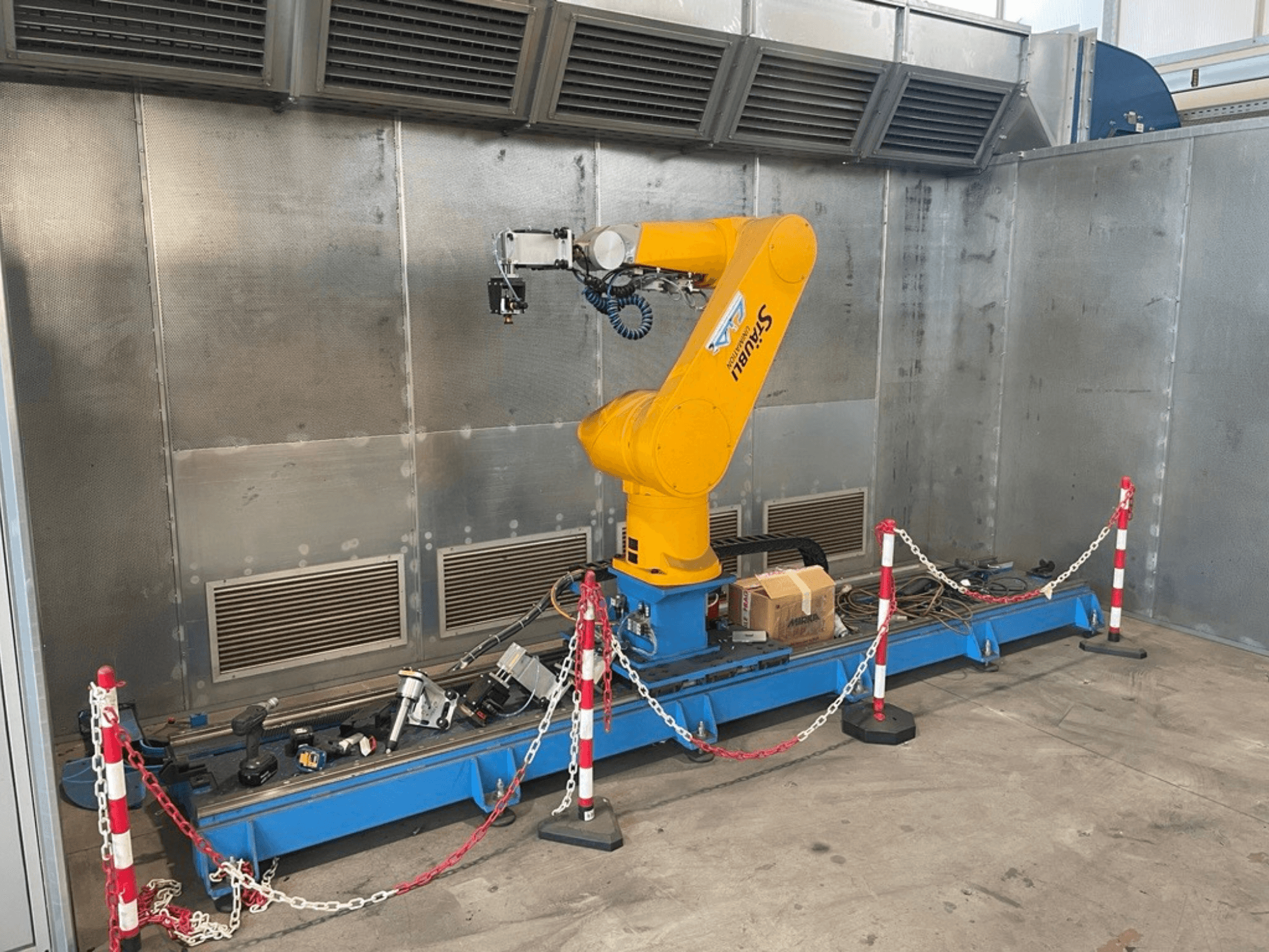 STAUBLI CS8C-RX160 robota roka darbnīcā, skats no sāniem, ar kabeļiem un dažādiem instrumentiem uz zilas pamatnes, ko ieskauj aizsargbarjeras.