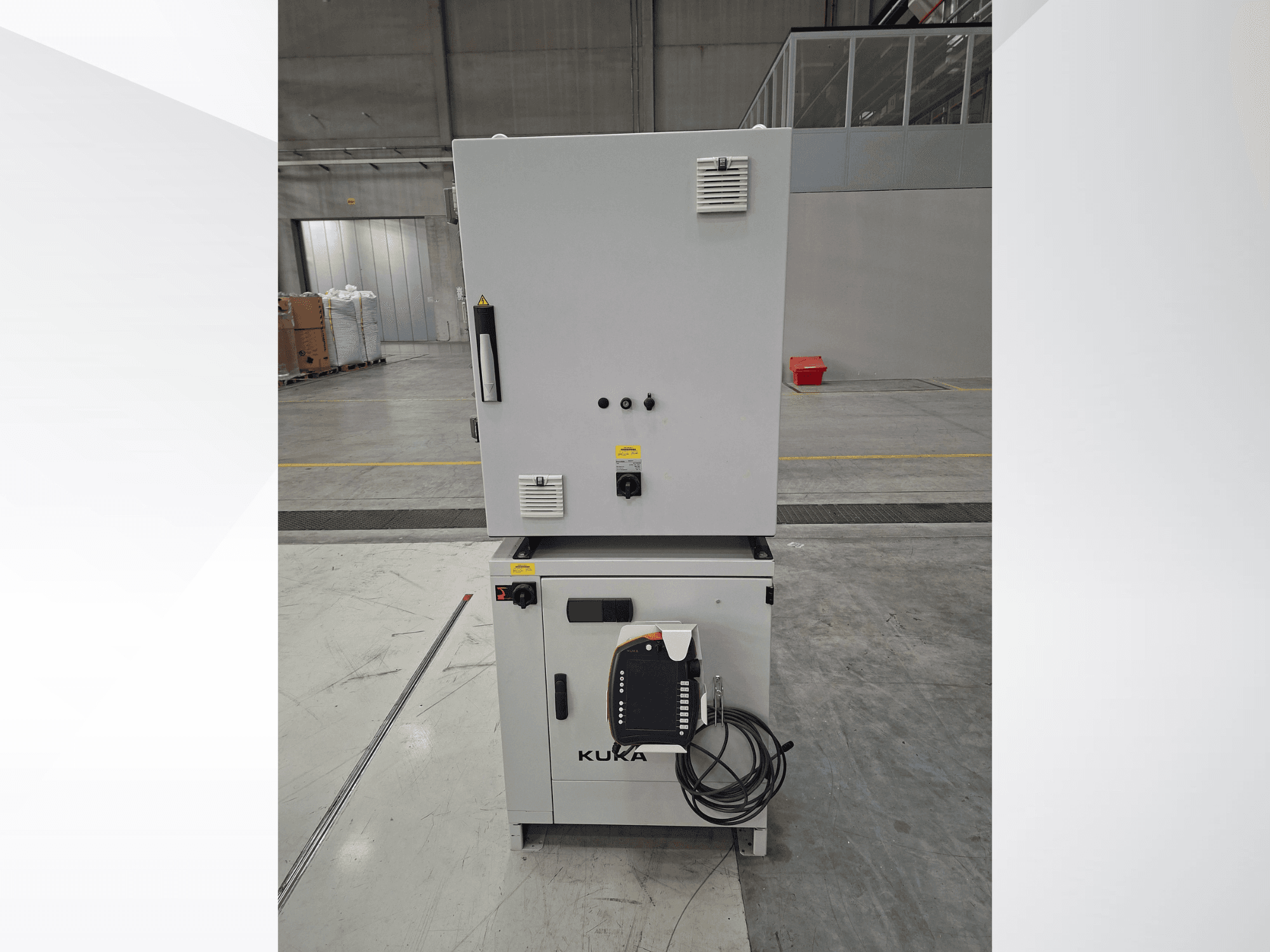 KUKA KR 90 R2700 PRO C4 FLR robota manipulatora vadības bloks — skats no priekšpuses, kurā redzams vadības panelis un drošības uzlīmes plašā noliktavā.
