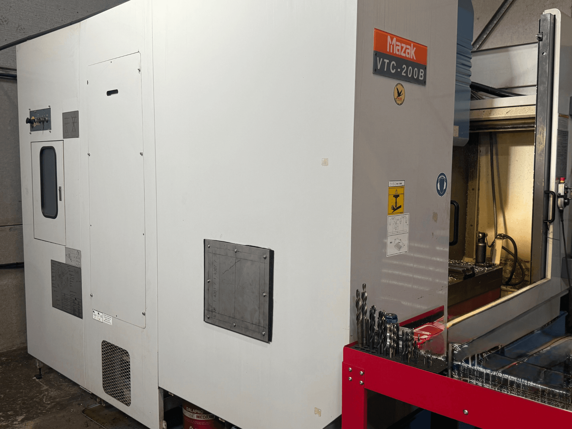 Mazak VTC-200B vertikālais apstrādes centrs no sāniem, kurā redzamas marķētas vadības ierīces un urbji uz sarkana uzglabāšanas plaukta.