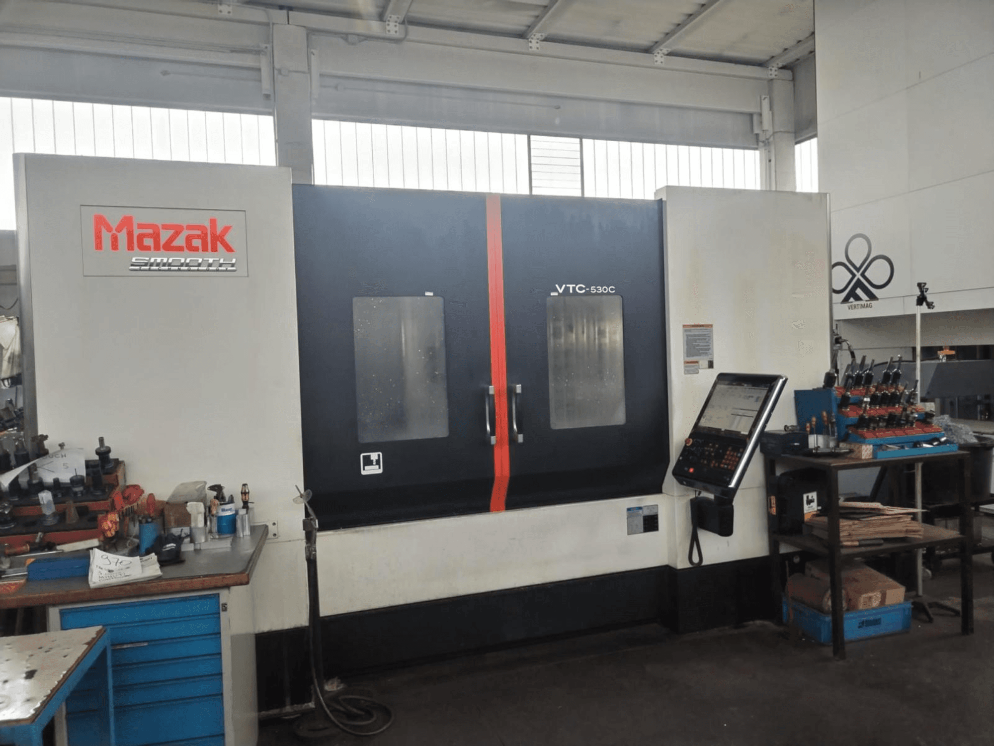 Vertikālais apstrādes centrs „Mazak VTC-530C“ darbnīcā, kurā redzams vadības panelis un instrumenti uz sānu galda.