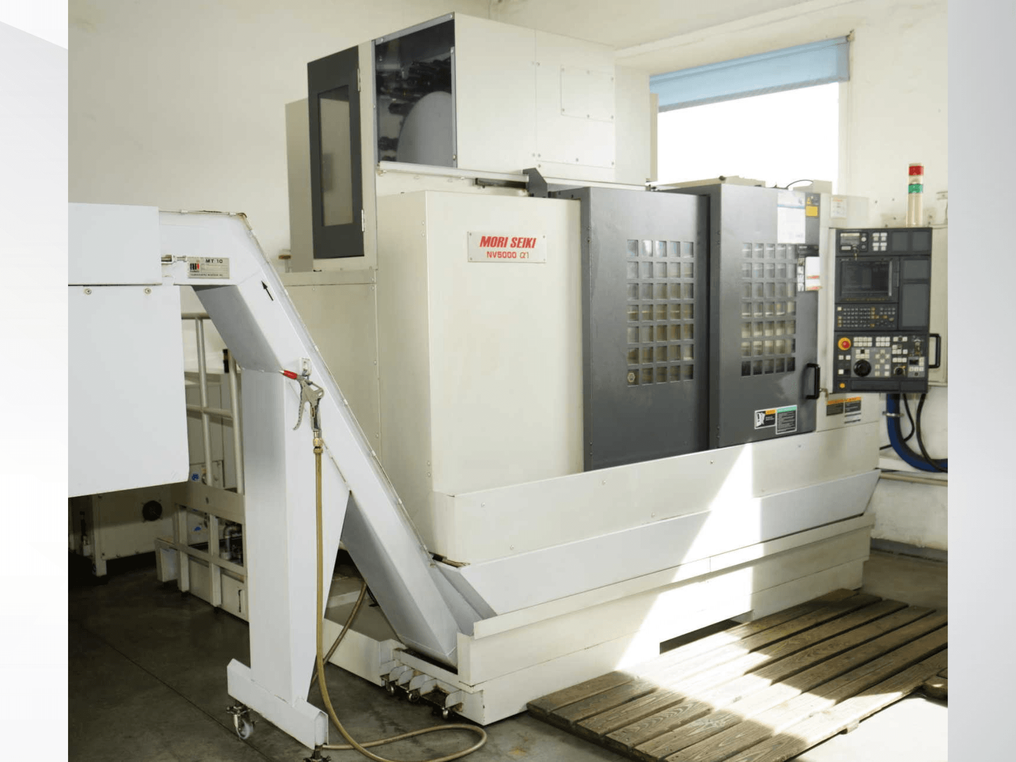 MORI SEIKI NV 5000 alpha1 vertikālais apstrādes centrs, skats no priekšpuses, kurā redzams vadības panelis, drošības elementi un iekraušanas zona.
