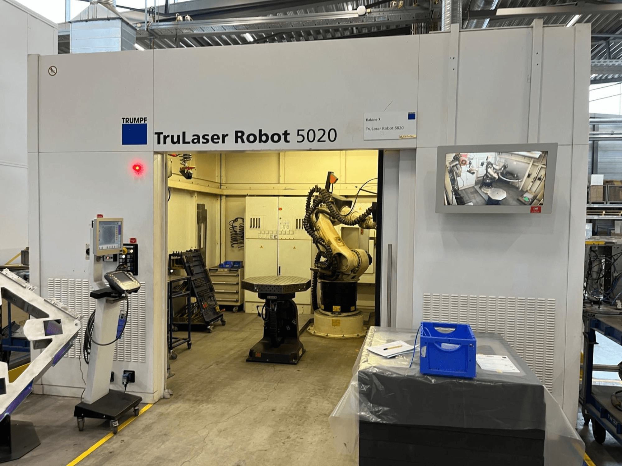 TRUMPF TruLaser Robot 5020 rūpnieciskā vidē, kurā redzama vadības pults, robota roka un monitors, uz kura tiek attēlotas darbības.