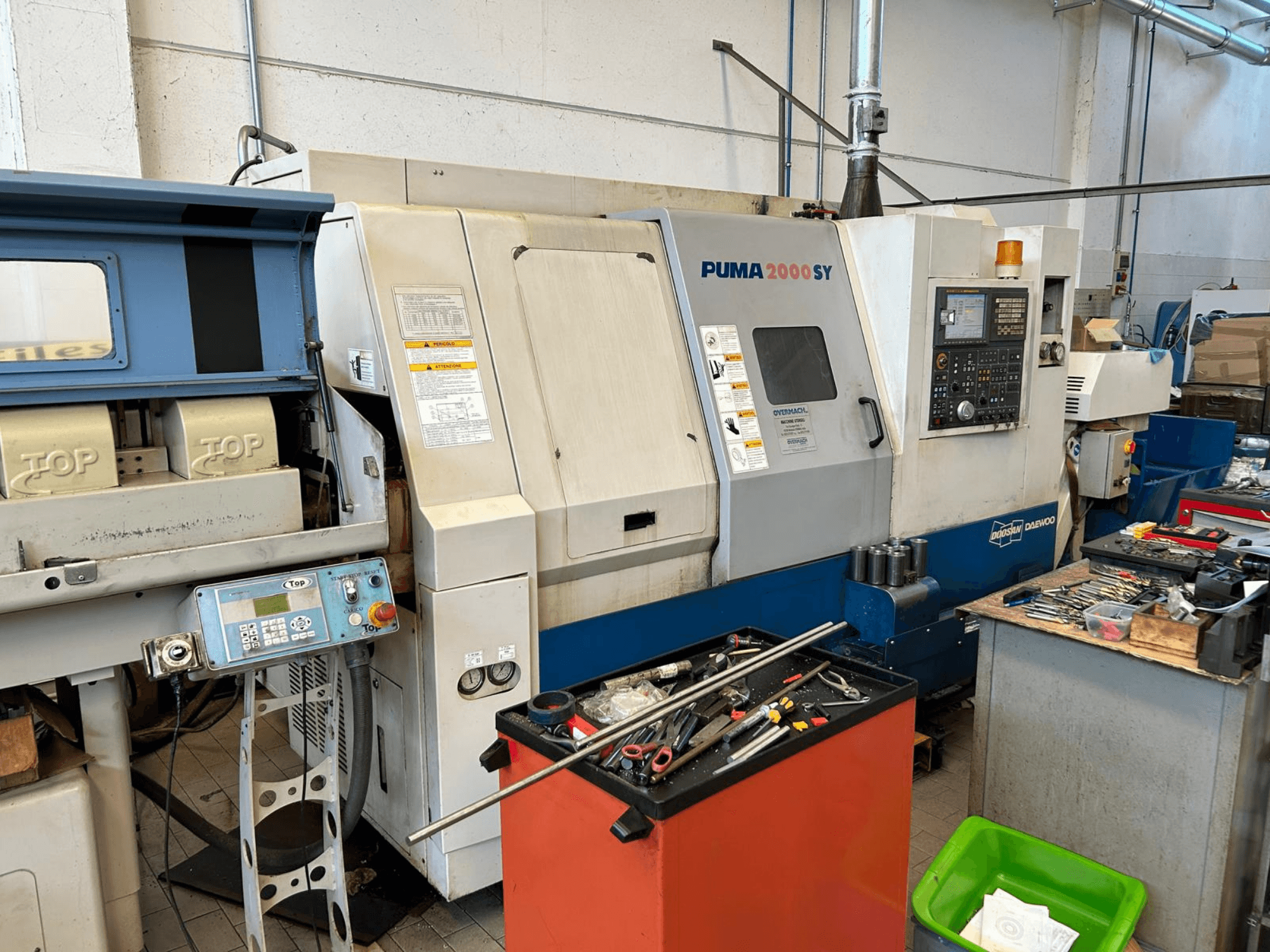 DOOSAN PUMA 2000SY horizontālā virpošanas mašīna darbnīcā, sānskats, kurā redzams vadības panelis un blakus esošie darbarīki.