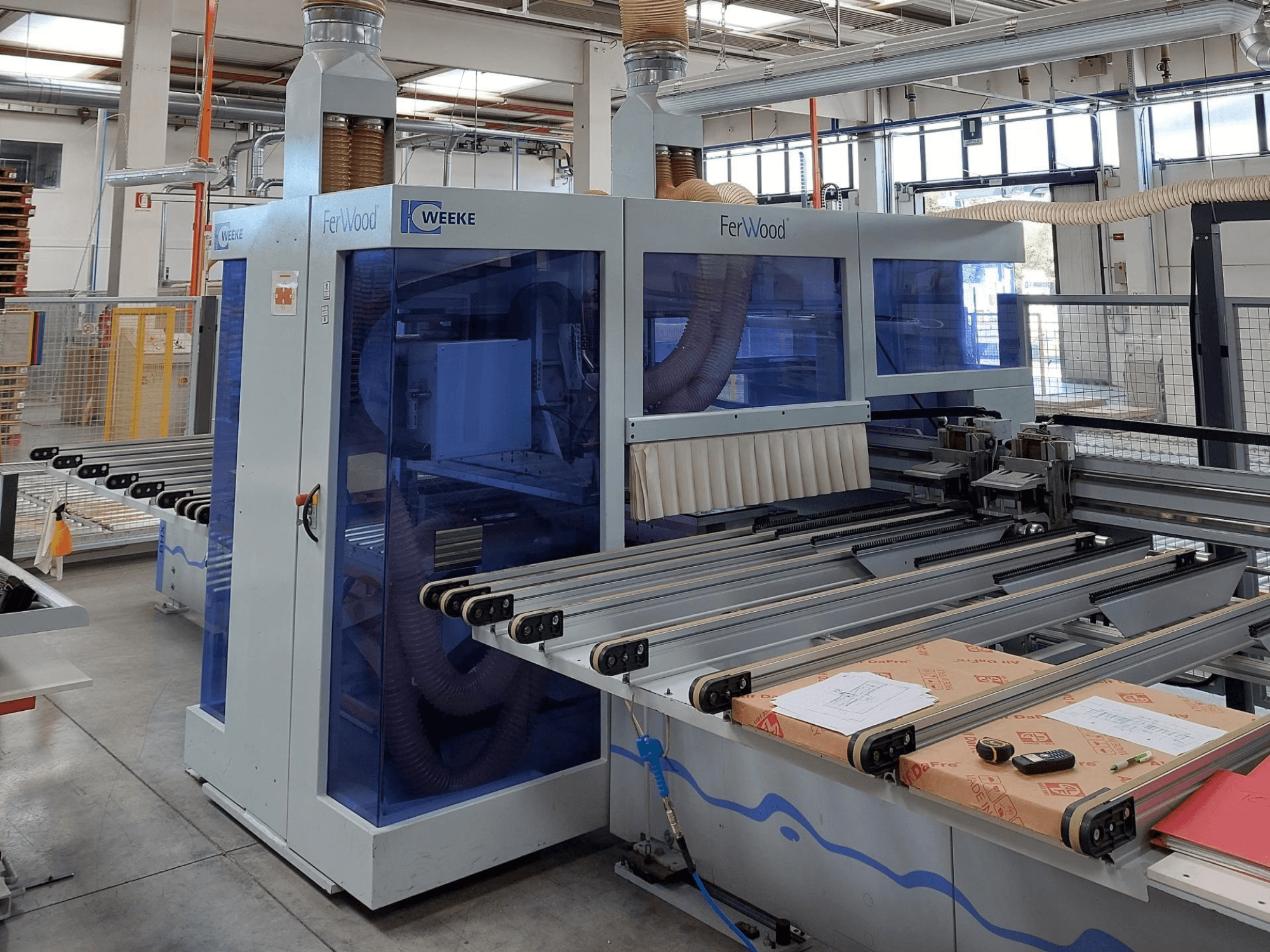 Weeke FerWood CNC mašīna darbnīcā, priekšējais skats, kurā redzami zili tonēti paneļi, vadības pogas un darba galds ar materiāliem.