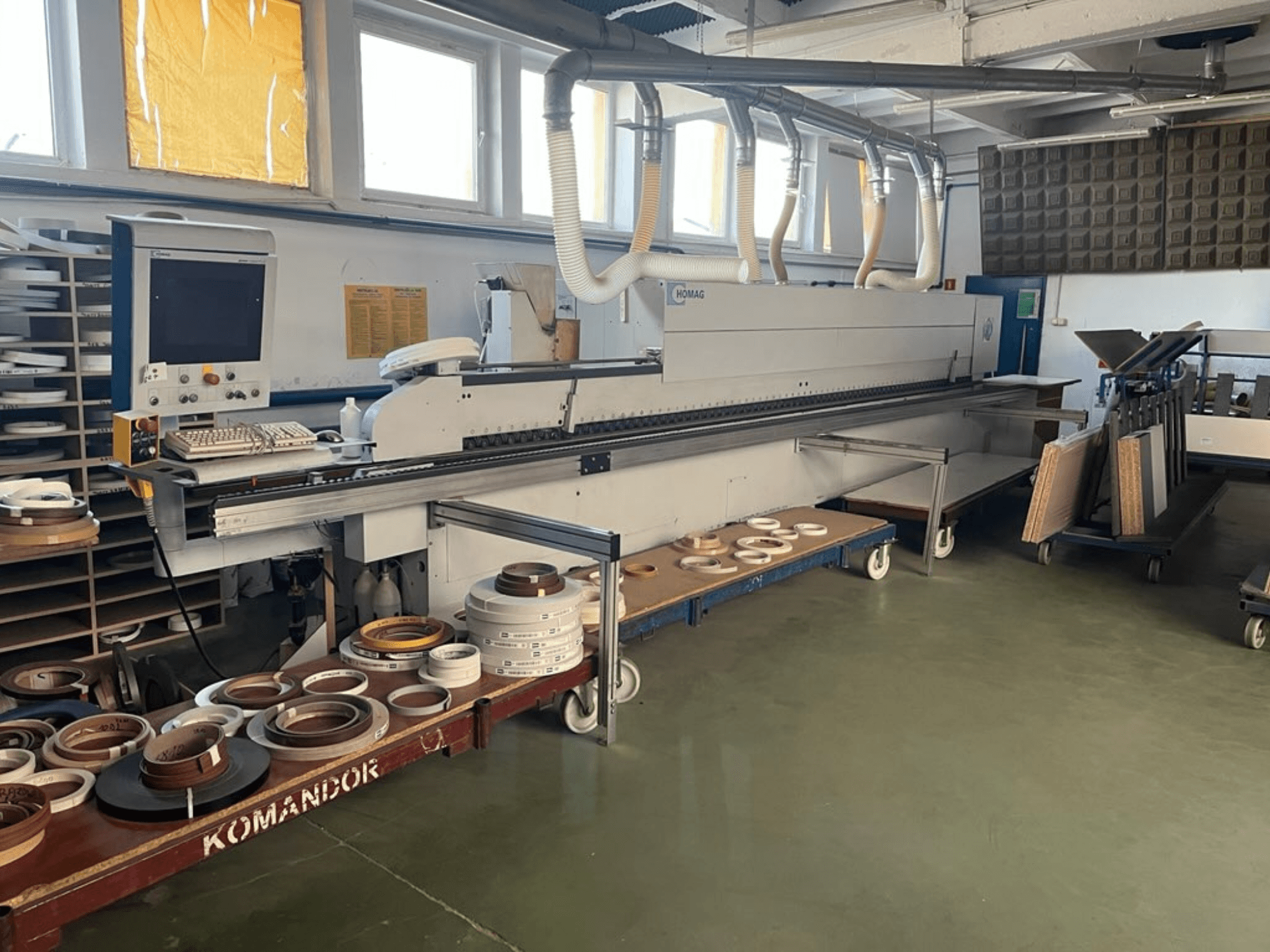 HOMAG malu aplīmēšanas iekārta darbnīcā, ar vadības ierīcēm un dažādiem malu aplīmēšanas materiāliem uz zila ratiņa. Skats no malas.