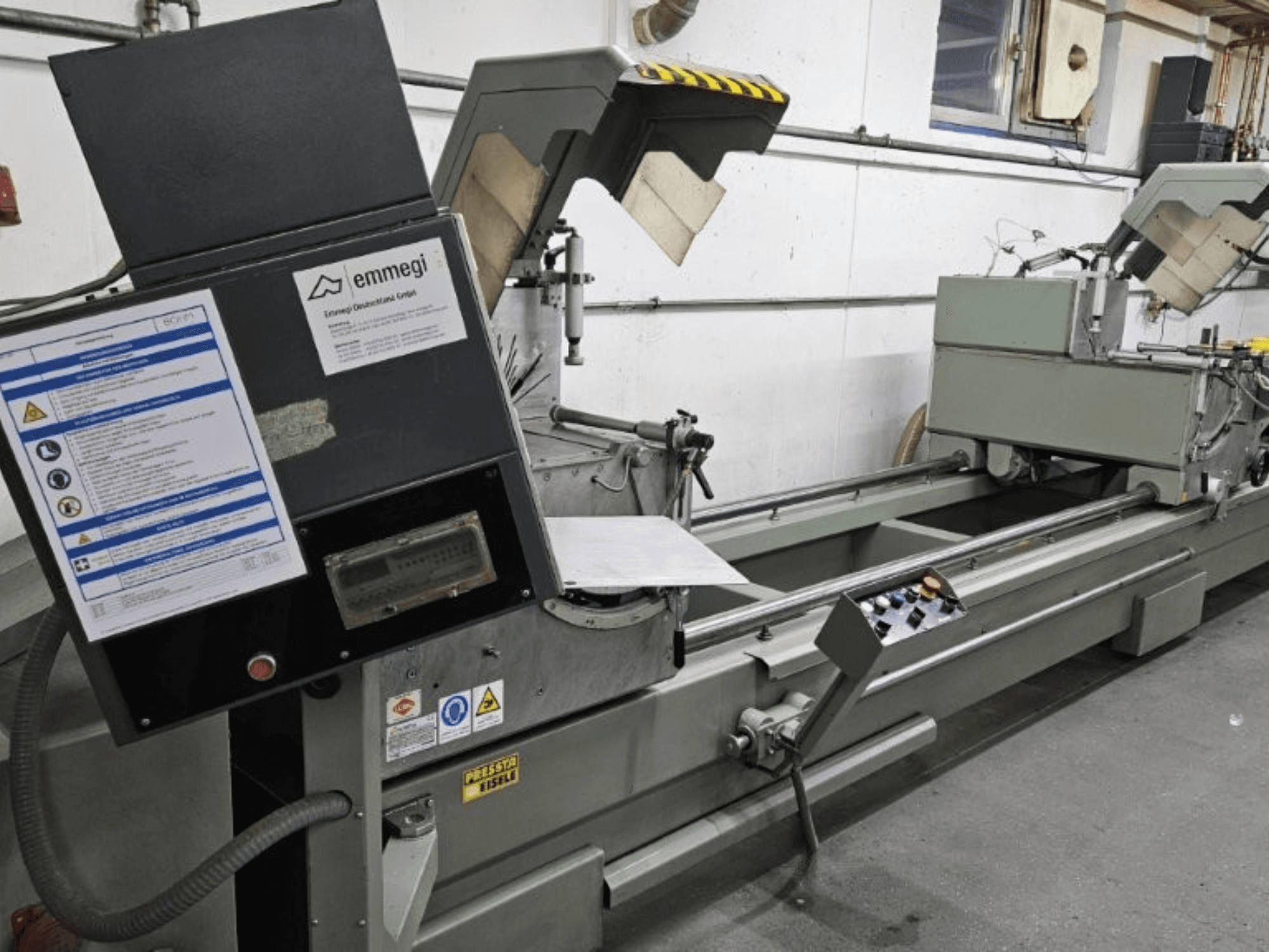 Emmegi rūpnieciskā griešanas iekārta, skats no priekšpuses, ar vadības paneli, drošības marķējumiem un darbības sastāvdaļām.