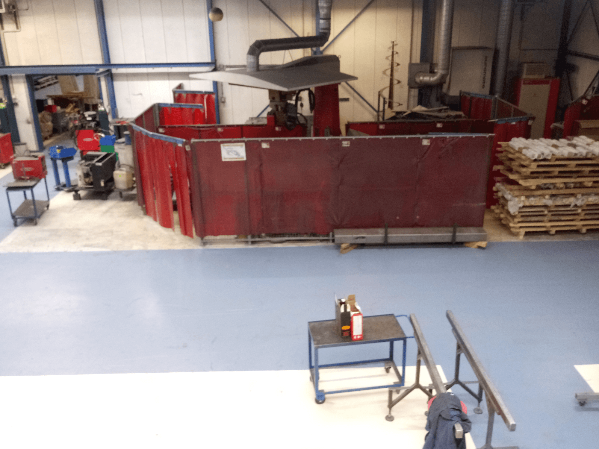 Rūpnieciskā darba telpa ar sarkaniem metināšanas aizkariem, mašīnām un paletēm; skats no augšas, kurā redzams izkārtojums un iekārtas.