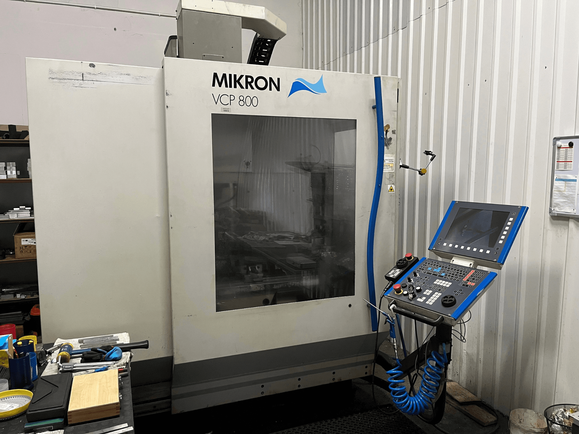 Vertikālais apstrādes centrs „MIKRON VCP 800“, skats no priekšpuses, priekšplānā redzams vadības panelis un darbarīki uz darba galda.