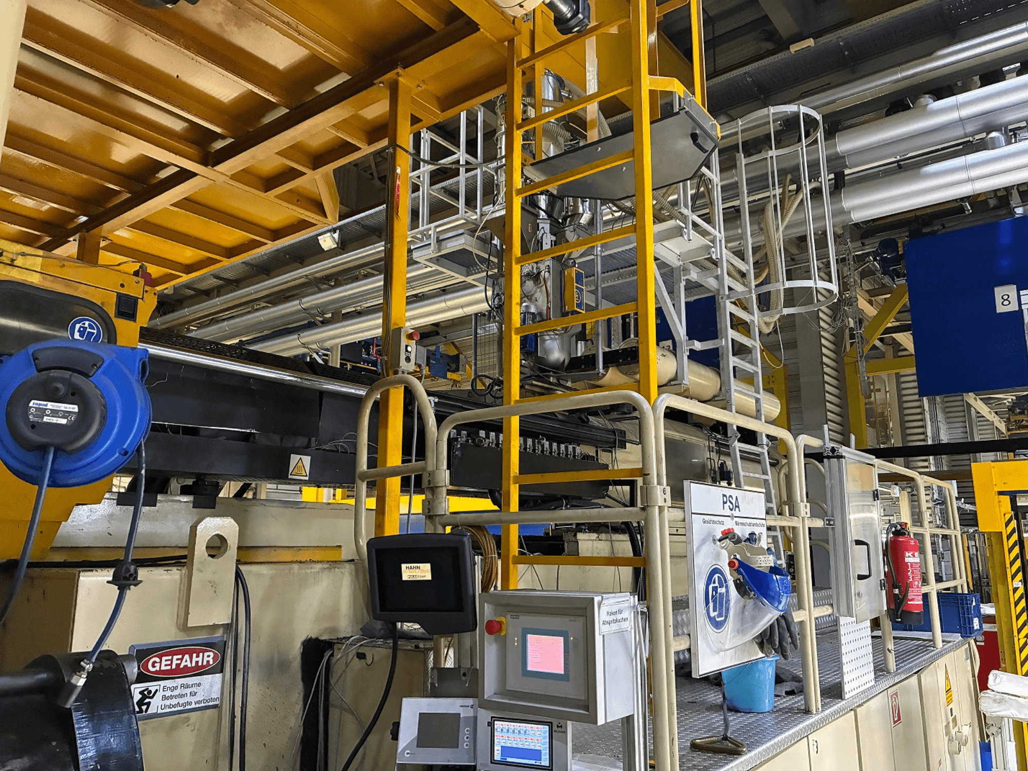 Rūpniecisko mašīnu vadības panelis ar zilu drošības aprīkojumu un dzeltenām sastatnēm, skats no malas, kurā redzamas caurules un brīdinājuma zīmes.