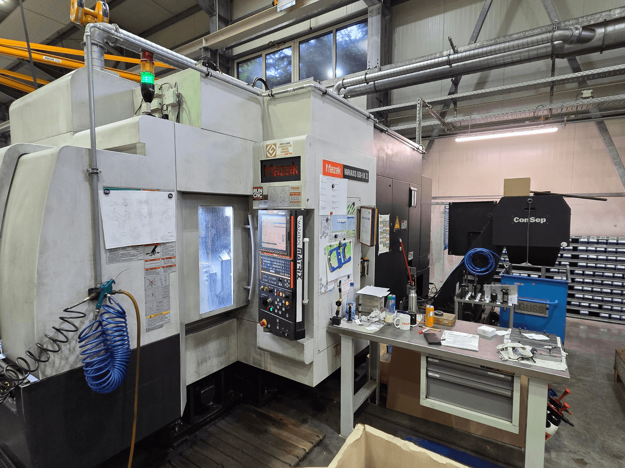 Mazak Variaxis 630-5X CNC darbnīcā, priekšējais skats ar vadības paneli, šļūteni un piederumiem uz sānu galda.