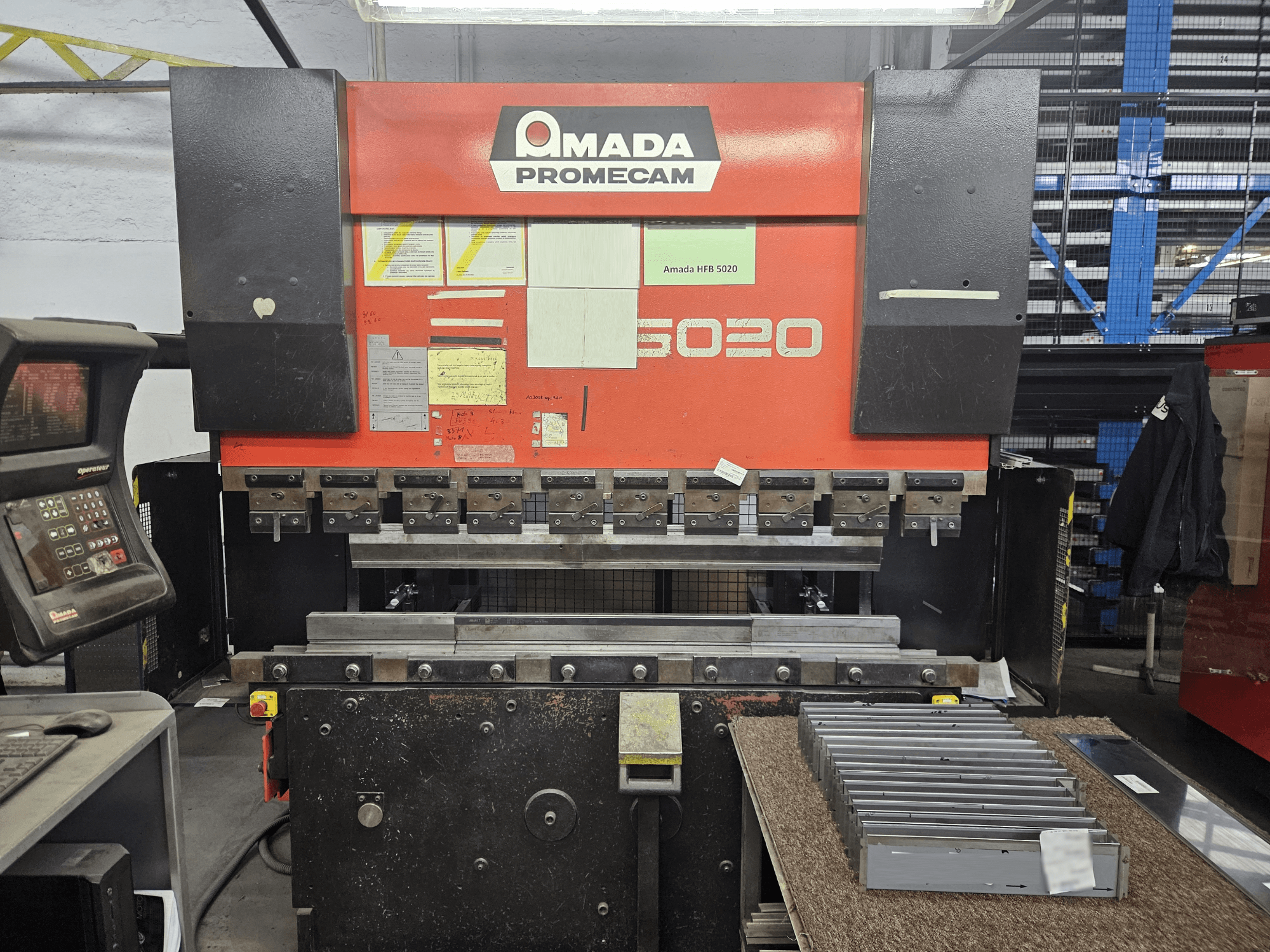 Amada HFBO 5020 preses bremze priekšpusē, ar vadības paneli un sakrautām metāla detaļām uz darba virsmas.