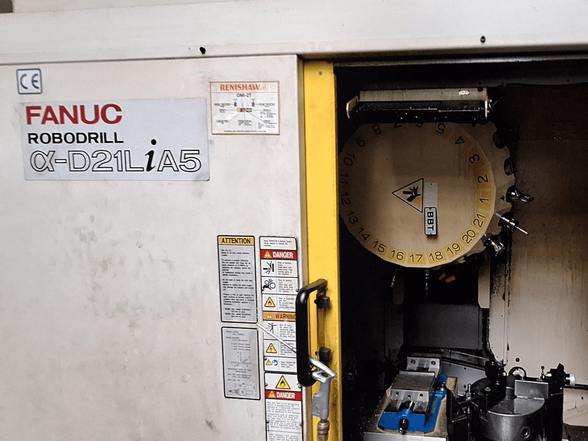 FANUC Robodrill α-D21LiA5, priekšējais skats. Parādīts instrumentu karuselis un apstrādes zona ar skavām un brīdinājumiem uz durvīm.