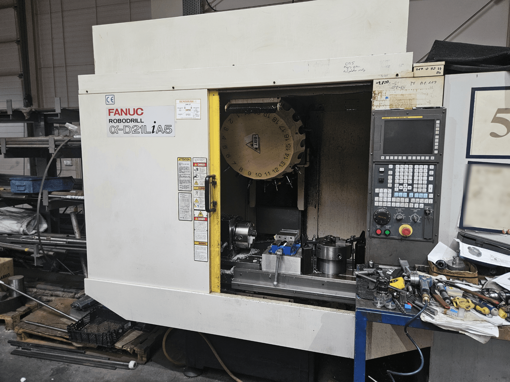 FANUC Robodrill α-D21LiA5, priekšējais skats, kurā redzams instrumentu turētājs un vadības panelis, ap kuru atrodas darbnīcas instrumenti un aprīkojums.