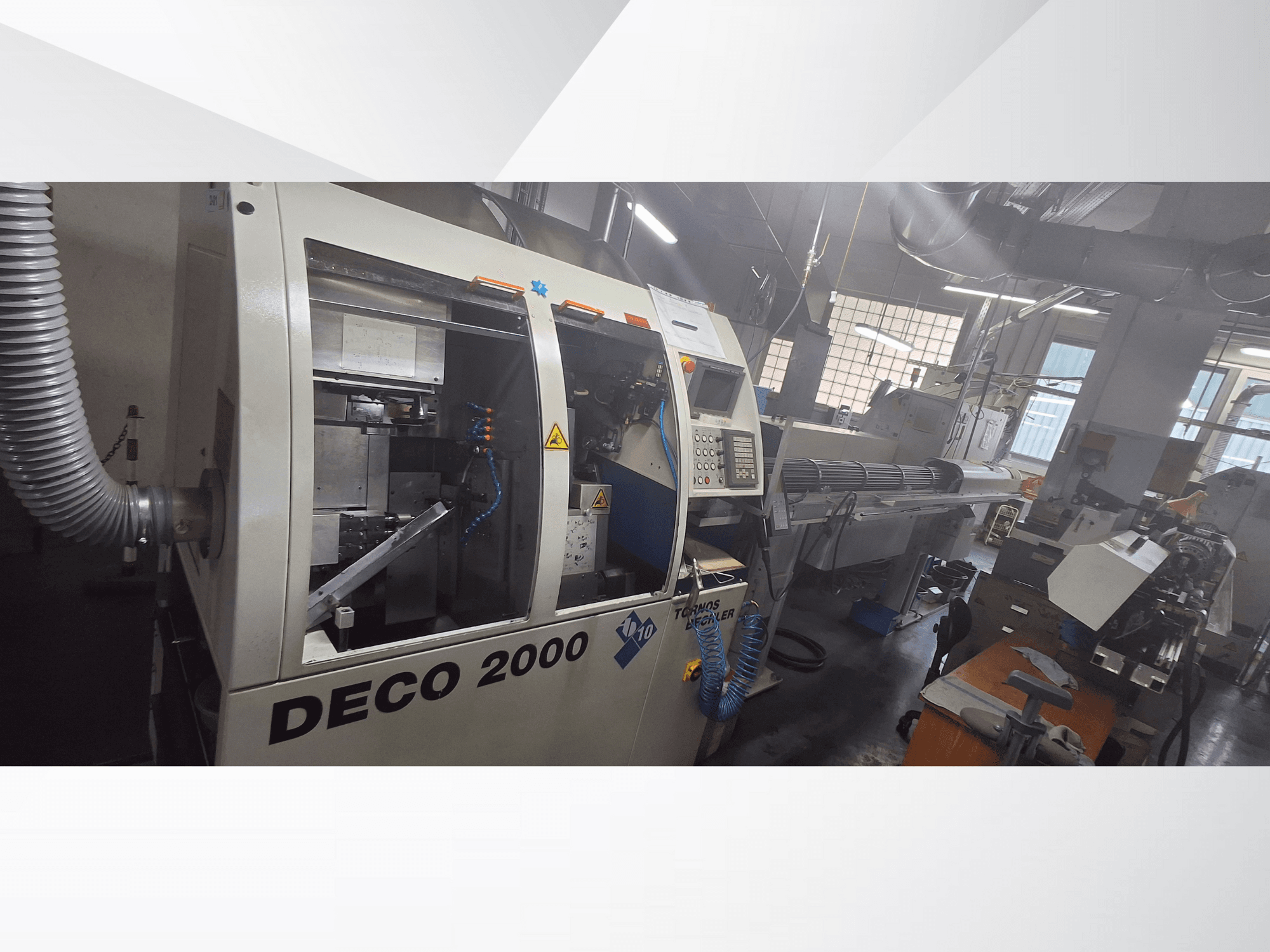 DECO 2000 CNC darbnīcā, skats no priekšpuses, redzamas vadības ierīces un garas sagataves uzstādīšana labajā pusē.