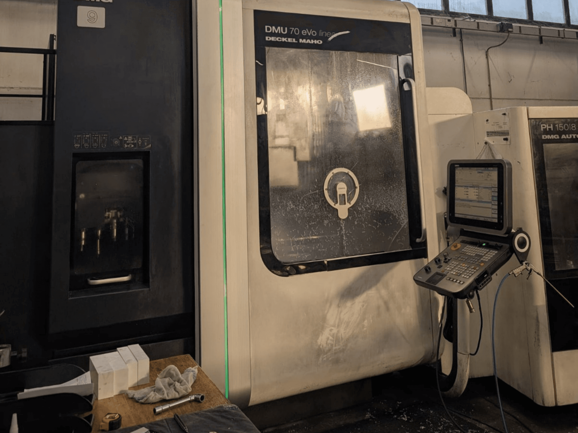 DMG DECKEL MAHO DMU 70 EVO Lineārais vertikālais apstrādes centrs, skats no priekšpuses ar redzamu vadības paneli, instrumentiem un darba vietu.