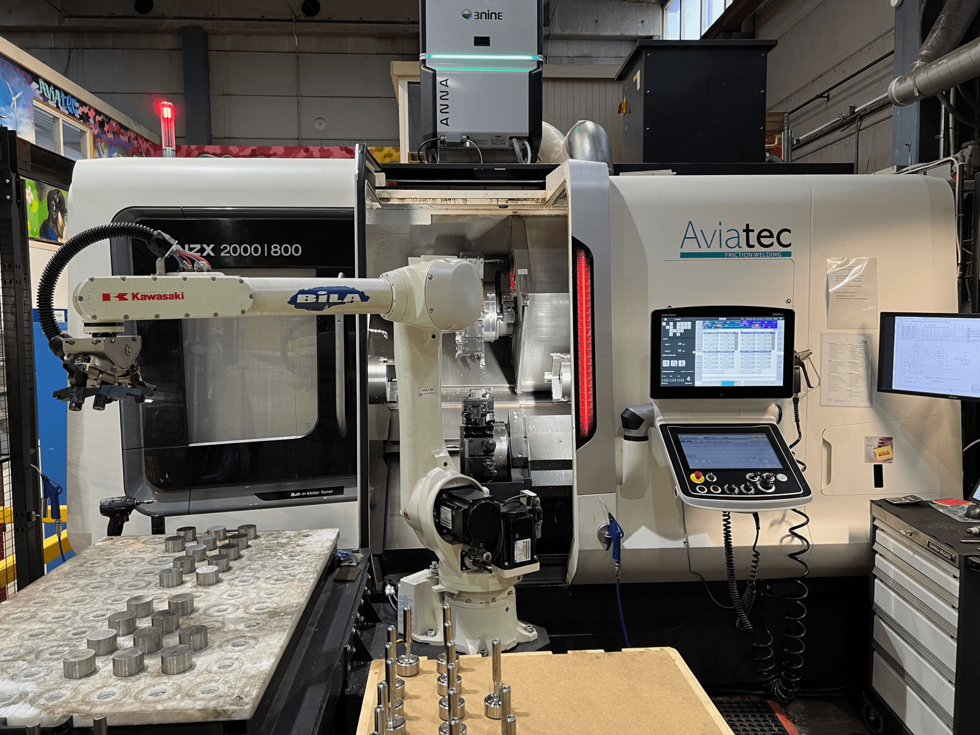 Horizontālā virpošanas mašīna DMG MORI NZX 2000/800SY ar „Kawasaki“ robotizēto roku, vadības pulti un metāla detaļām uz darba galda.