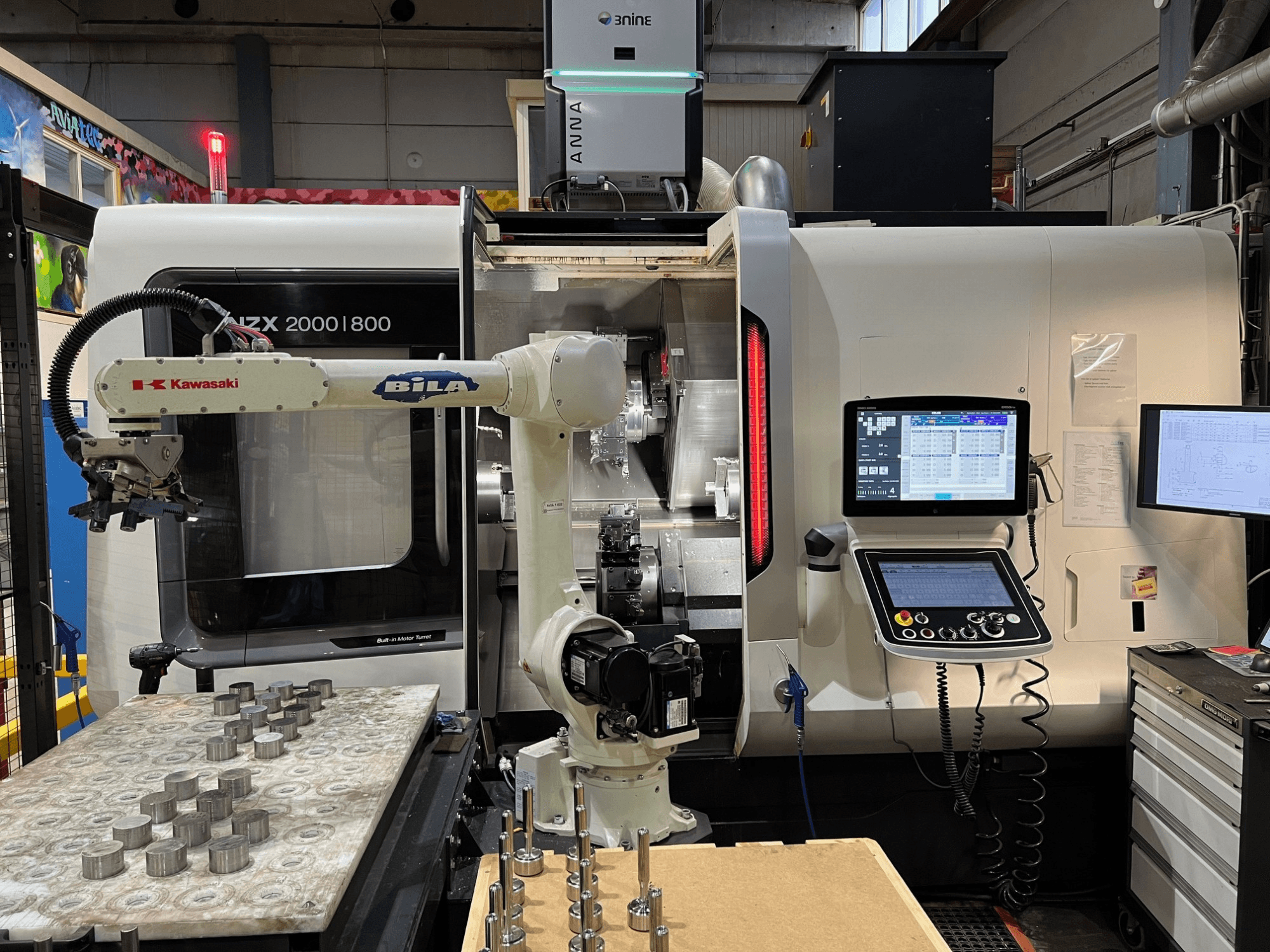 Horizontālā virpošanas mašīna DMG MORI NZX 2000/800SY darbnīcā, kur redzama „Kawasaki“ robotizētā roka un vadības pultis.