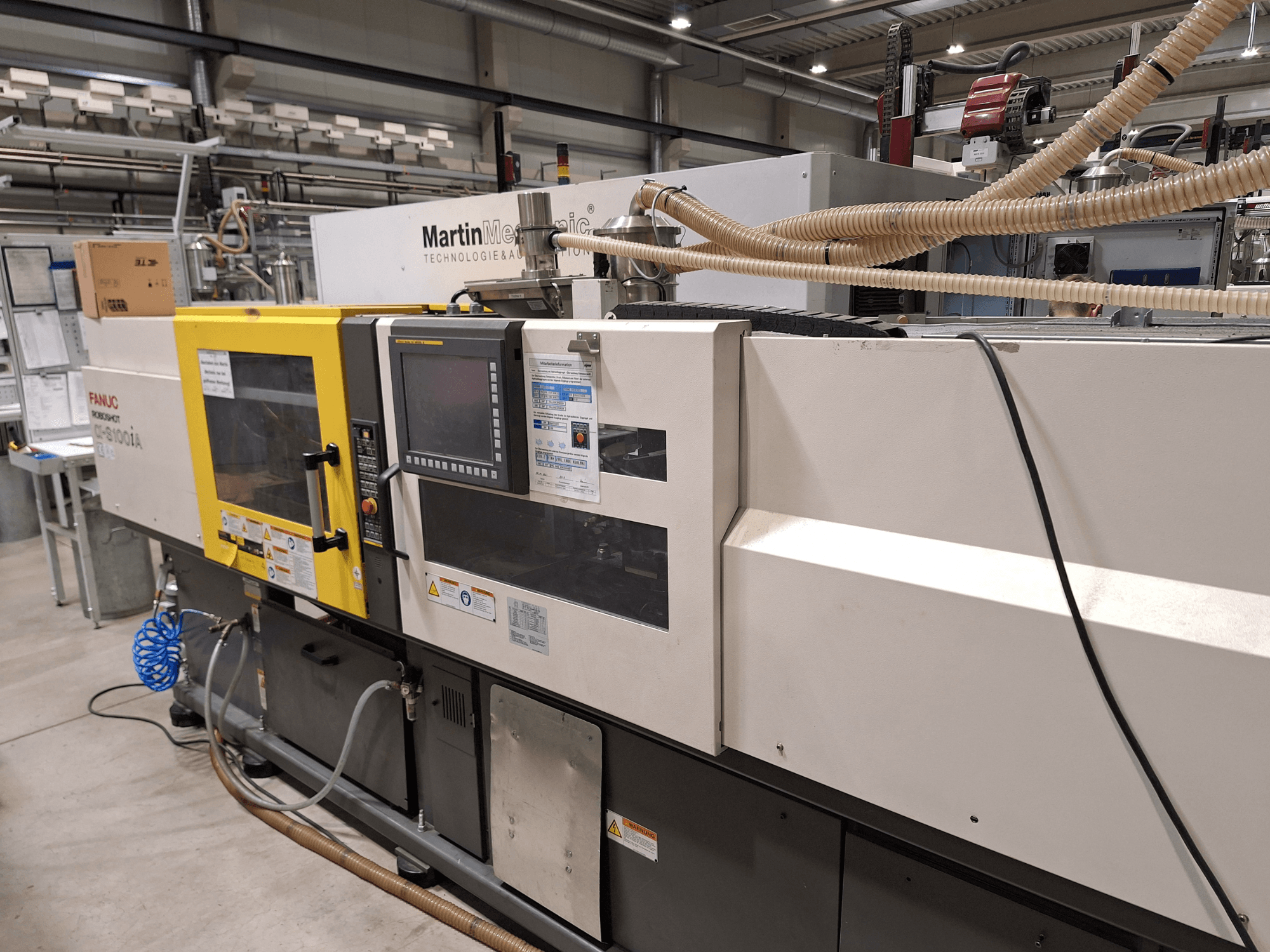 FANUC Roboshot S100iA elektriskā iesmidzināšanas iekārta, skats no sāniem, aprīkota ar skārienjūtīgo ekrānu un drošības marķējumu.