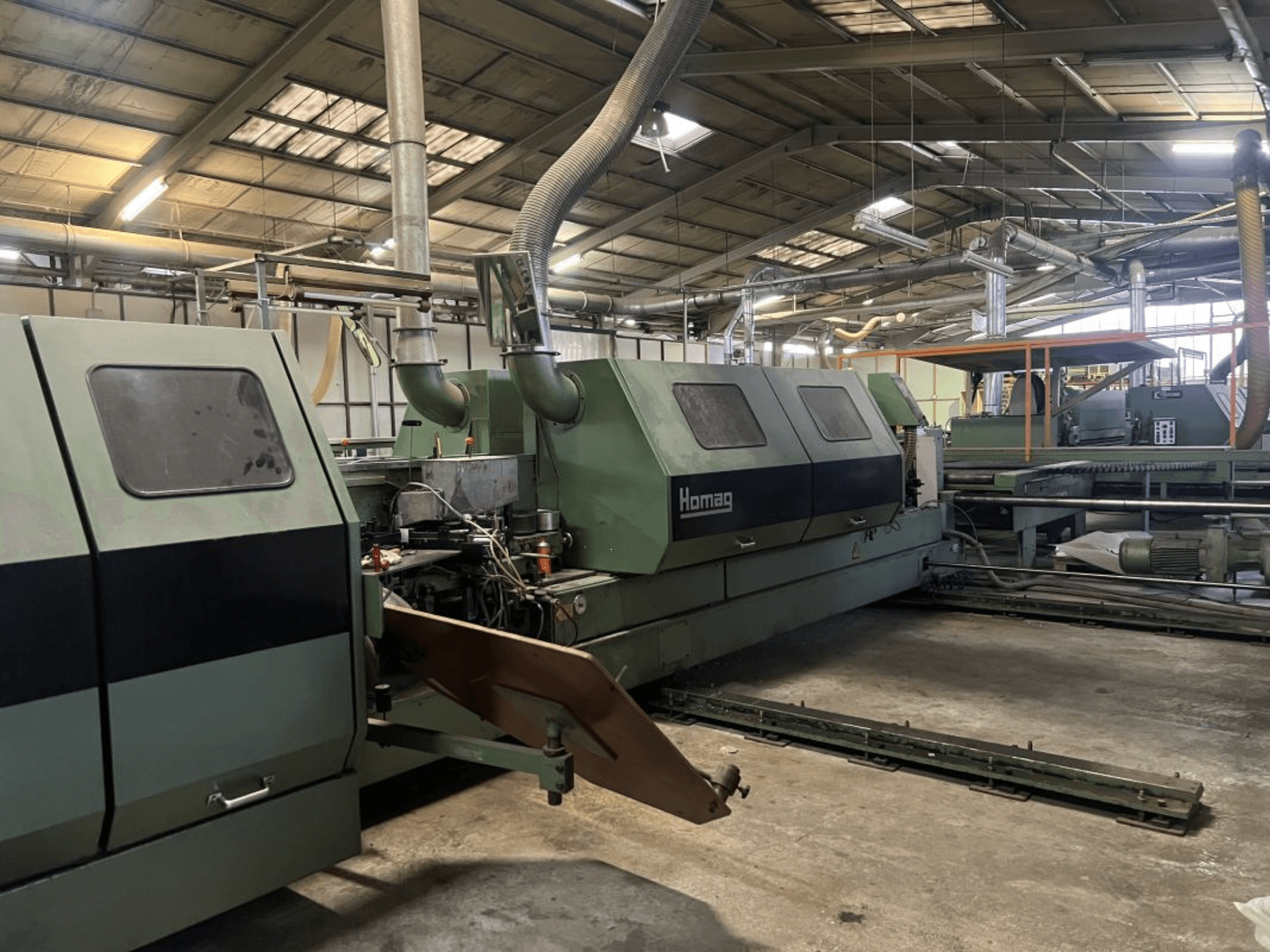 Homag industriālā mašīna zaļā krāsā, skats no sāniem, ar redzamām darbības sastāvdaļām un vadības paneli noliktavā.