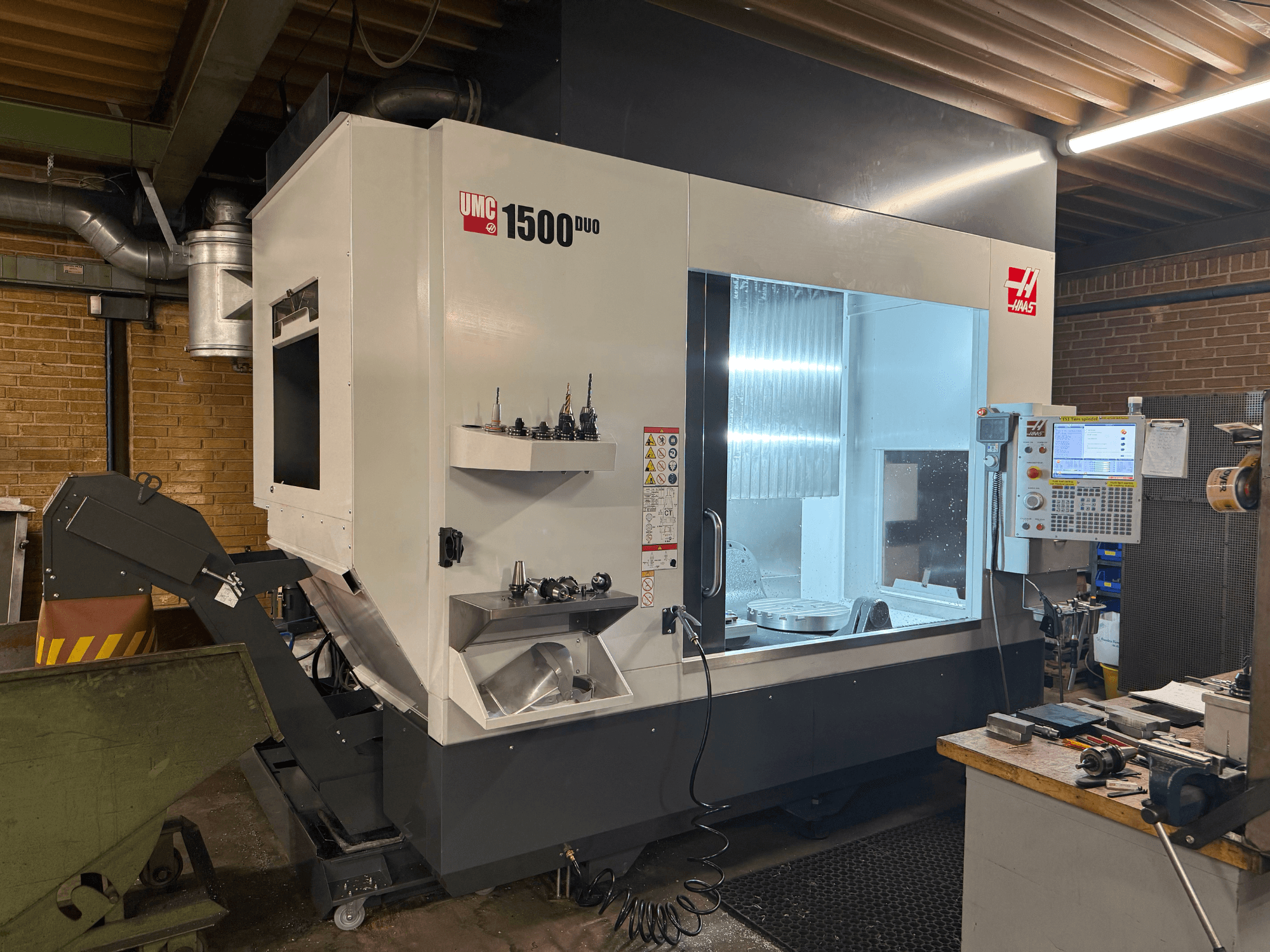 Haas UMC 1500DUO CNC darbgalds, skats no priekšpuses, ar lielu logu, vadības paneli un instrumentu turētājiem uz plaukta.