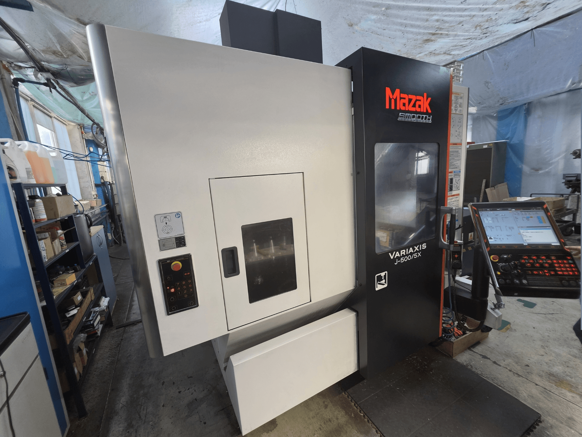 Mazak VARIAXIS J-500/5X vertikālais apstrādes centrs, skats no priekšpuses, ar vadības paneli un lielu apstrādes nodalījuma logu.