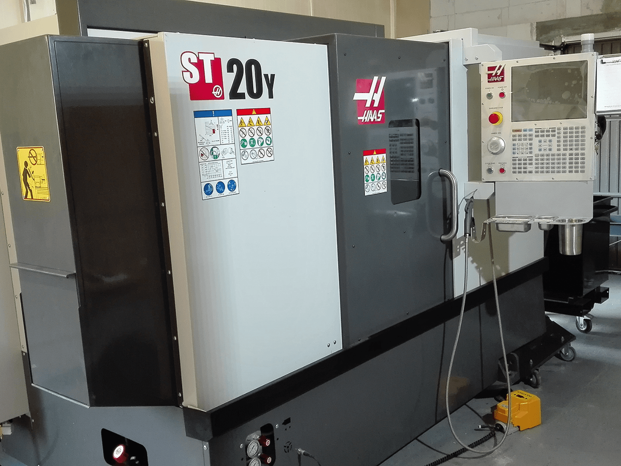 Haas ST20Y CNC virpa priekšpusē, ar vadības paneli, drošības marķējumiem un dzesēšanas šķidruma sistēmu.