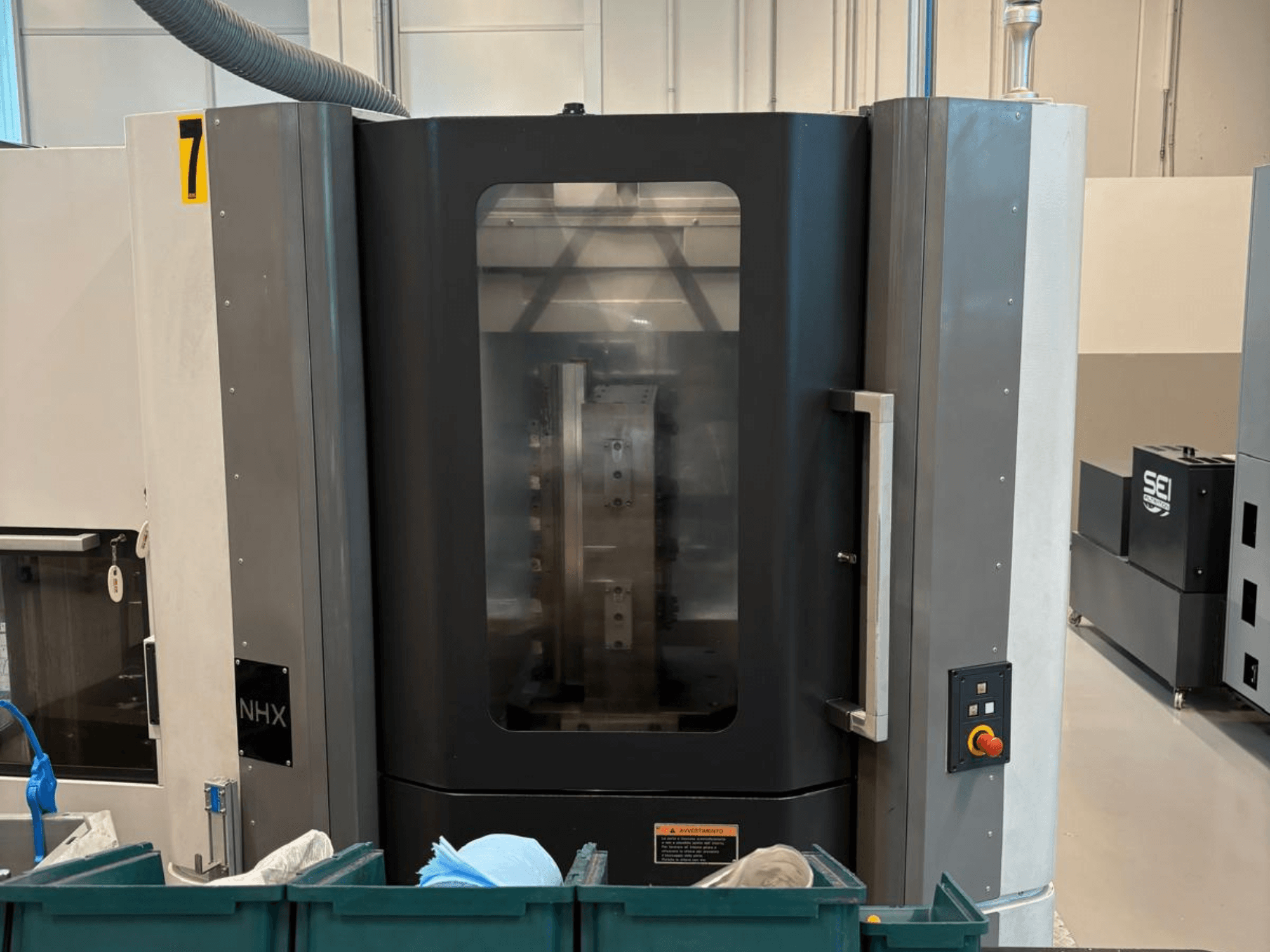 NHX CNC darbgalds priekšpusē, redzams vadības panelis un pārredzams displejs; atrodas darbnīcā ar zaļām tvertnēm blakus.