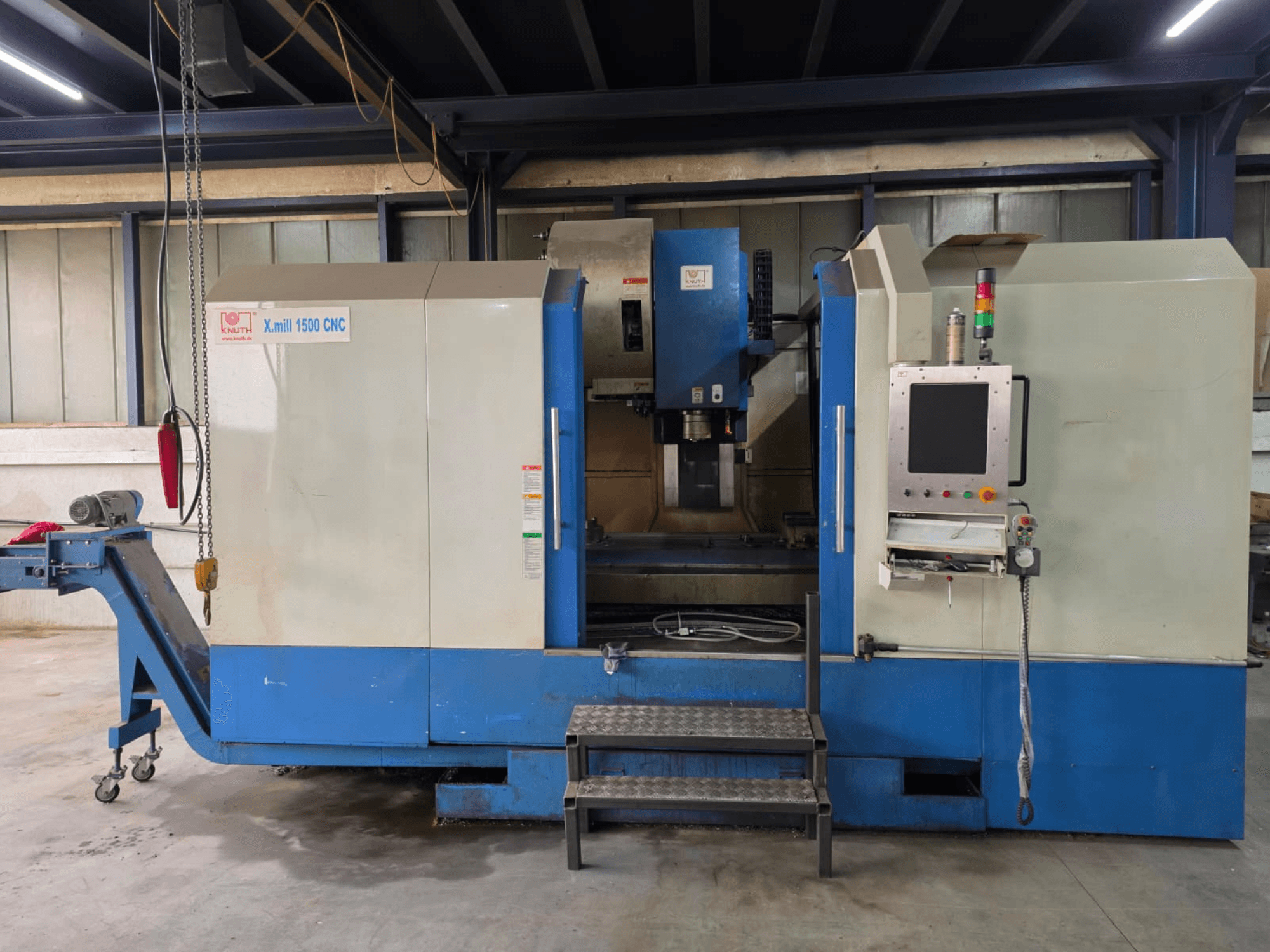 CNC vertikālais apstrādes centrs „KNUTH X-MILL 1500“, skats no sāniem; redzams vadības panelis, pakāpieni un izvades konveijers.