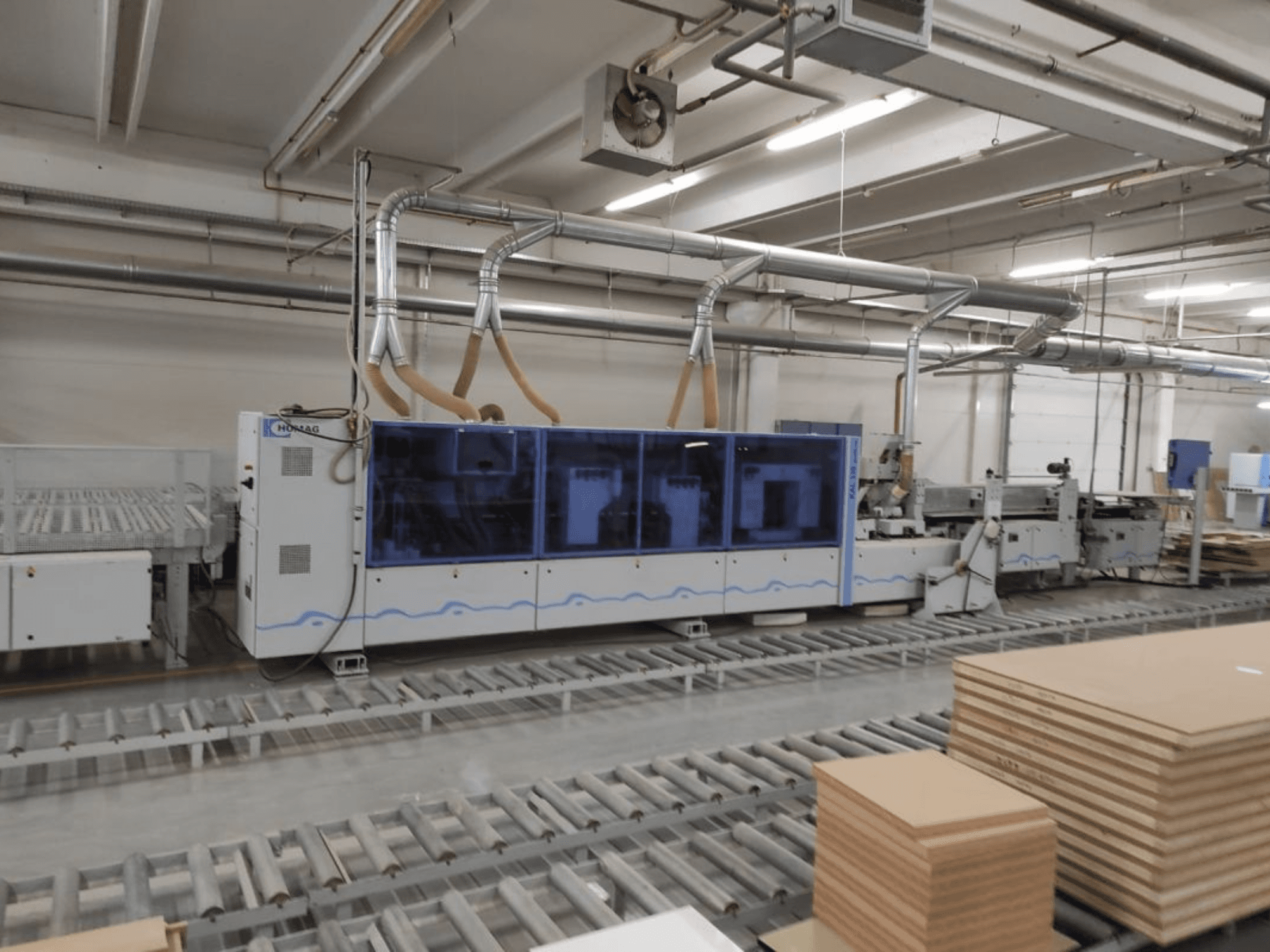 HOMAG ražošanas iekārta rūpnīcā, skats no priekšpuses, ar zilu korpusu un konveijera sistēmu ar sakrautām saplākšņa loksnēm.