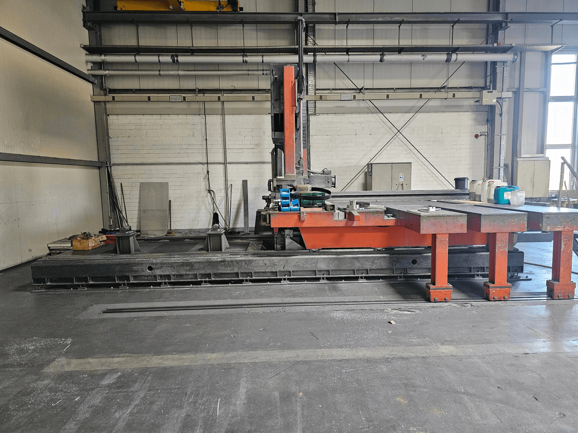 Rūpnieciskā griešanas mašīna atklātā darbnīcā, oranžā un melnā krāsā, ar lielu galdu; skats no sāniem, kurā redzami piederumi un darba vieta.