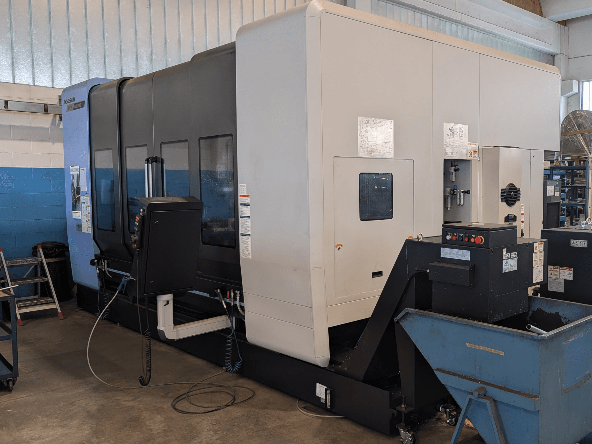 DOOSAN VCF850LSR-II vertikālais apstrādes centrs, skats no sāniem, kurā redzams vadības panelis, dzesēšanas šķidruma sistēma un izturīgā konstrukcija.