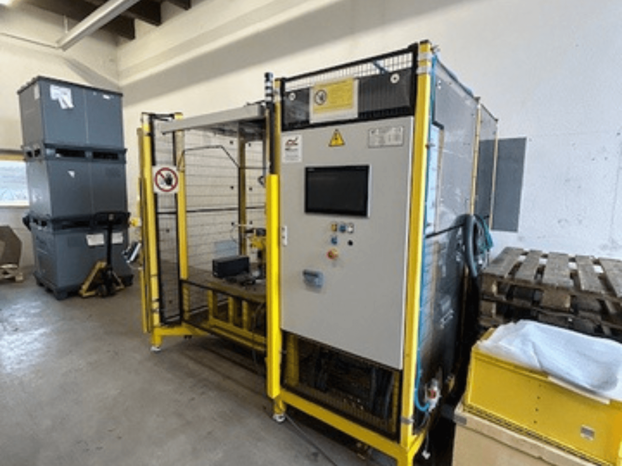 Rūpnieciskā mašīna ar vadības paneli un drošības elementiem, skats no priekšpuses; dzeltens rāmis, pelēkas tvertnes un paletes fonā.