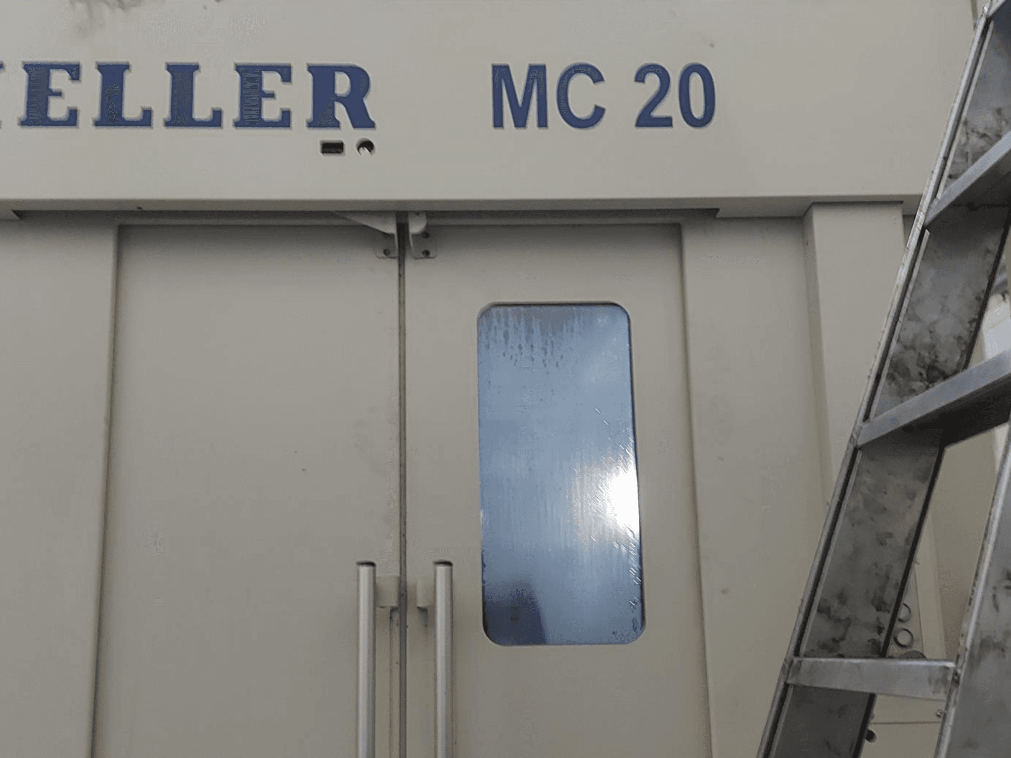 HELLER MC 20 horizontālā apstrādes centra priekšējais skats, redzami durvju rokturi un neliels logs. Kāpnes sānos.