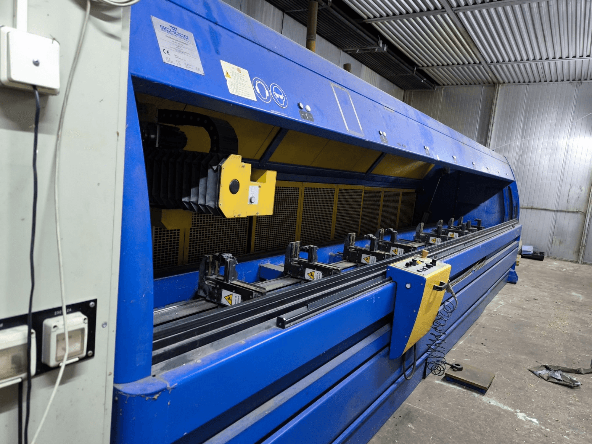 Schneider rūpnieciskā griešanas iekārta zilā krāsā, skats no sāniem, redzams vadības panelis, siksnas un vairākas fiksācijas stacijas.