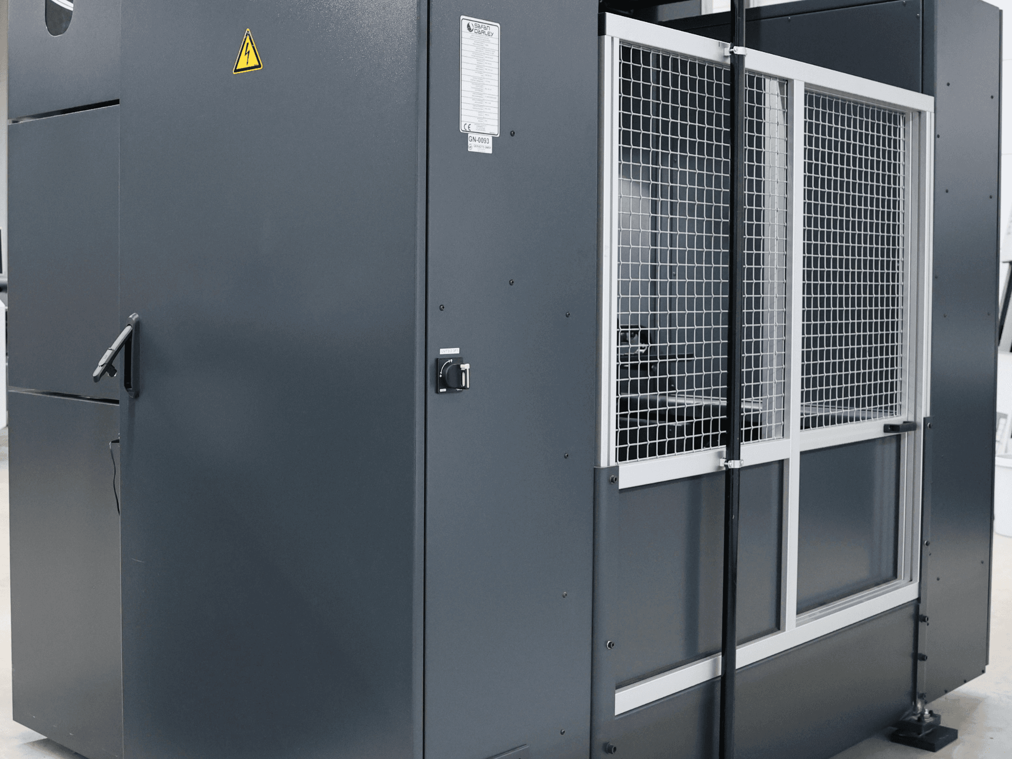 Pelēka rūpnieciska mašīna ar drošības brīdinājumu, priekšējais skats, kurā redzamas režģa durvis un vadības panelis, marķēts modelis.