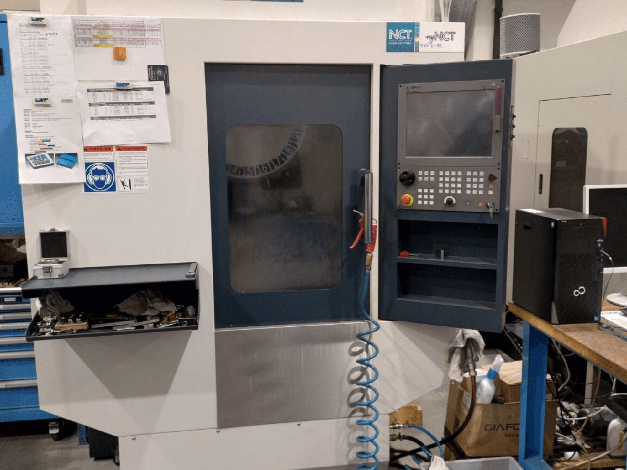 NCT CNC darbgalds, skats no priekšpuses, vadības panelis ar ciparu tastatūru, instrumenti glabāšanas atvilktnē un rokasgrāmata uz sienas.