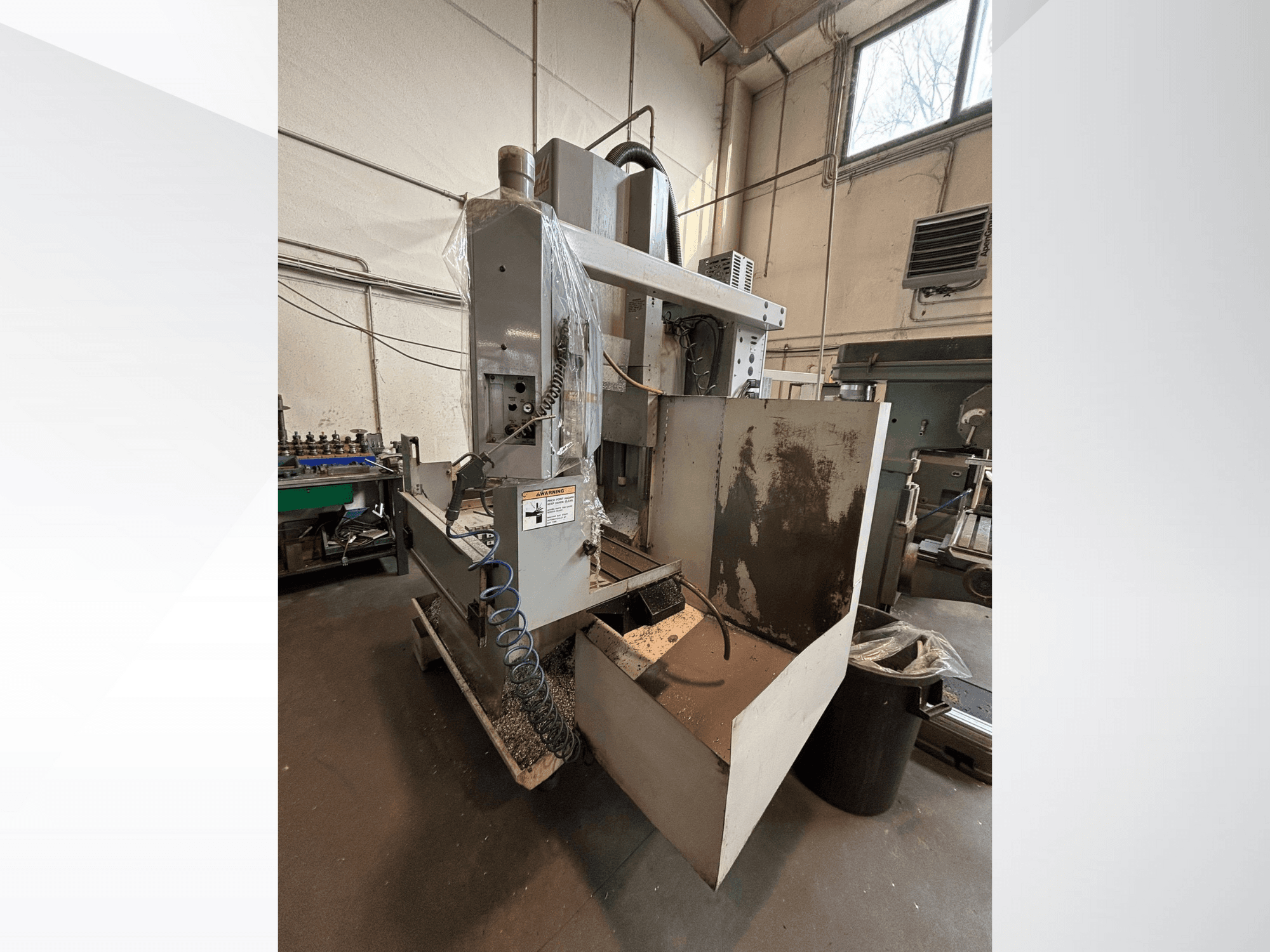 Vertikālā frēzmašīna ar aizsargvāciņu, skats no sāniem; redzama dzesēšanas šķidruma šļūtene un vadības panelis. Ideāli piemērots apstrādei.