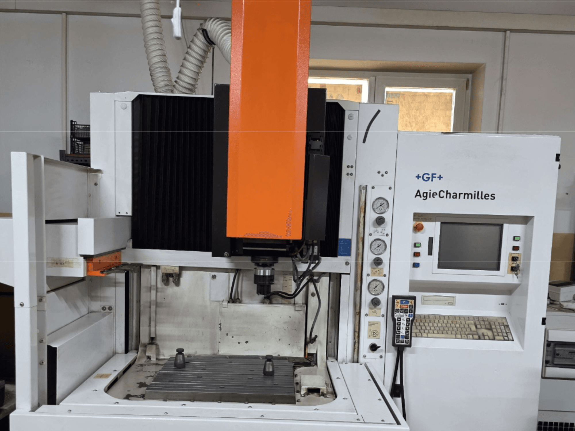 AgieCharmilles apstrādes centrs priekšpusē ar lielu oranžu vārpstu, vadības paneli un redzamu darba galdu.