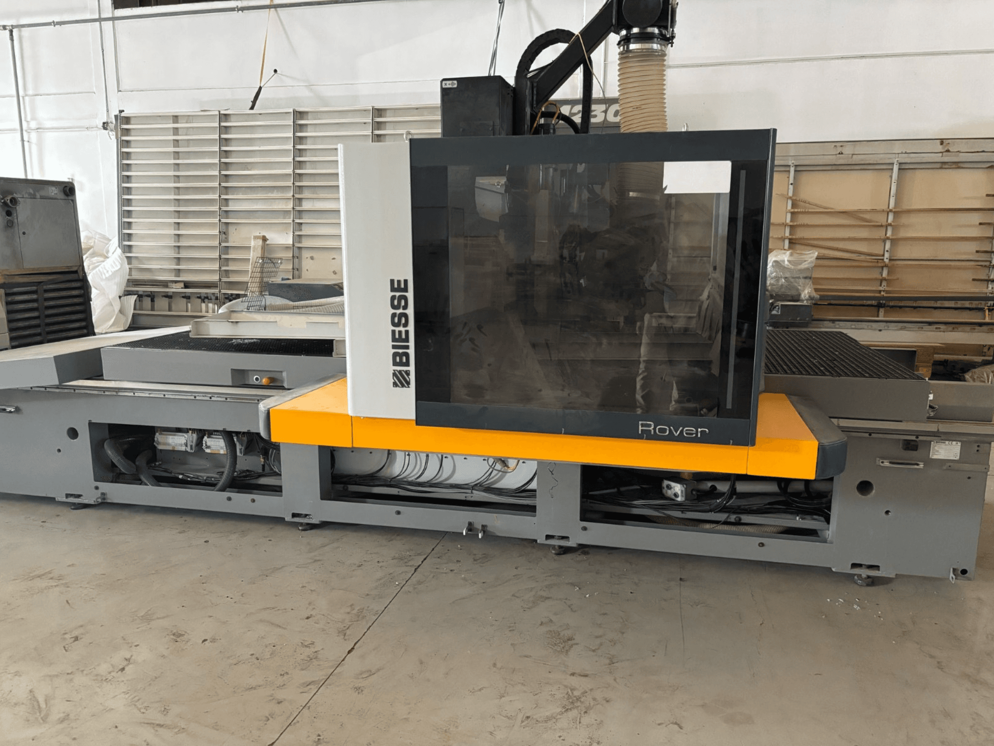 Biesse Rover CNC apstrādes centrs, skats no priekšpuses, ar lielu vadības paneli, dzelteniem akcentiem un atklātām mehāniskām detaļām.