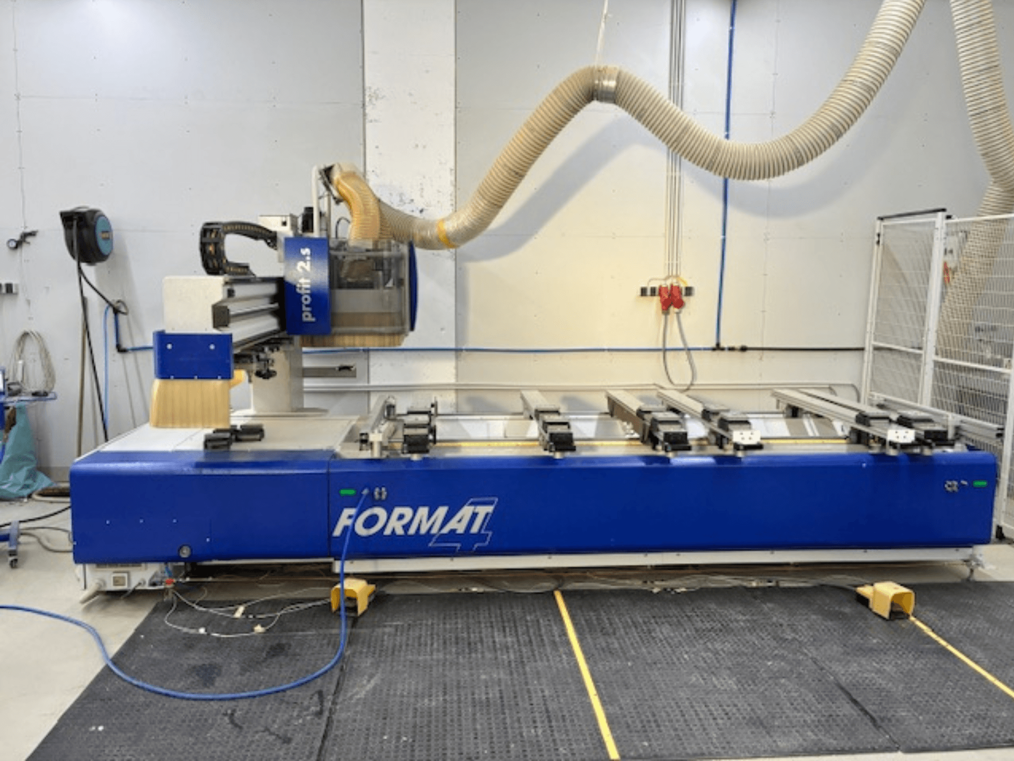 Zilais FORMAT 4 CNC frēzētājs, modelis Profit 2.5, sānu skats, kurā redzama griešanas zona un putekļu nosūces sistēma, uz gumijas paklāja.