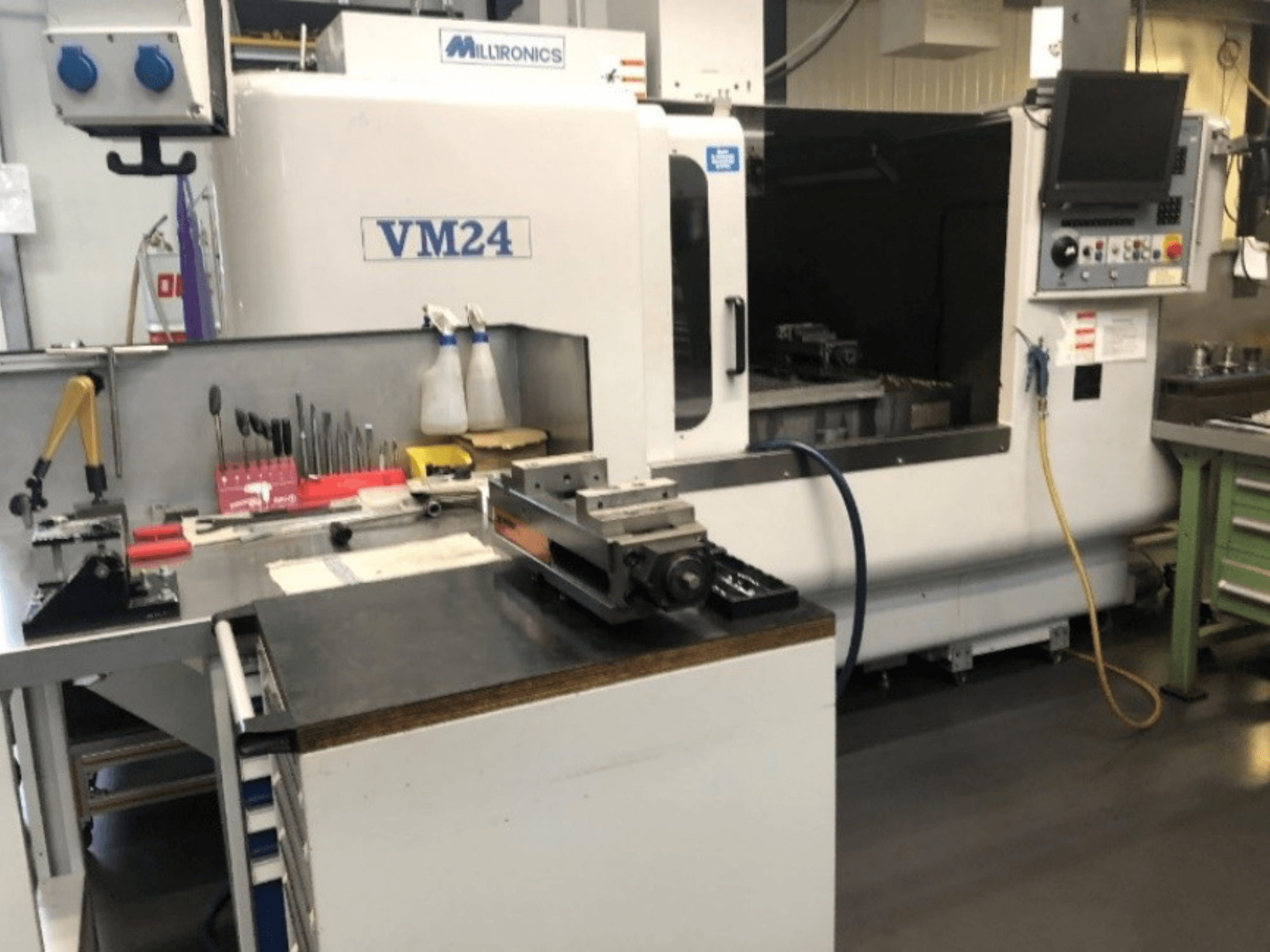 Mašīnu darba vieta ar Milltronics VM24 CNC mašīnu. Skats no priekšpuses ar instrumentiem un aprīkojumu uz darba galda.