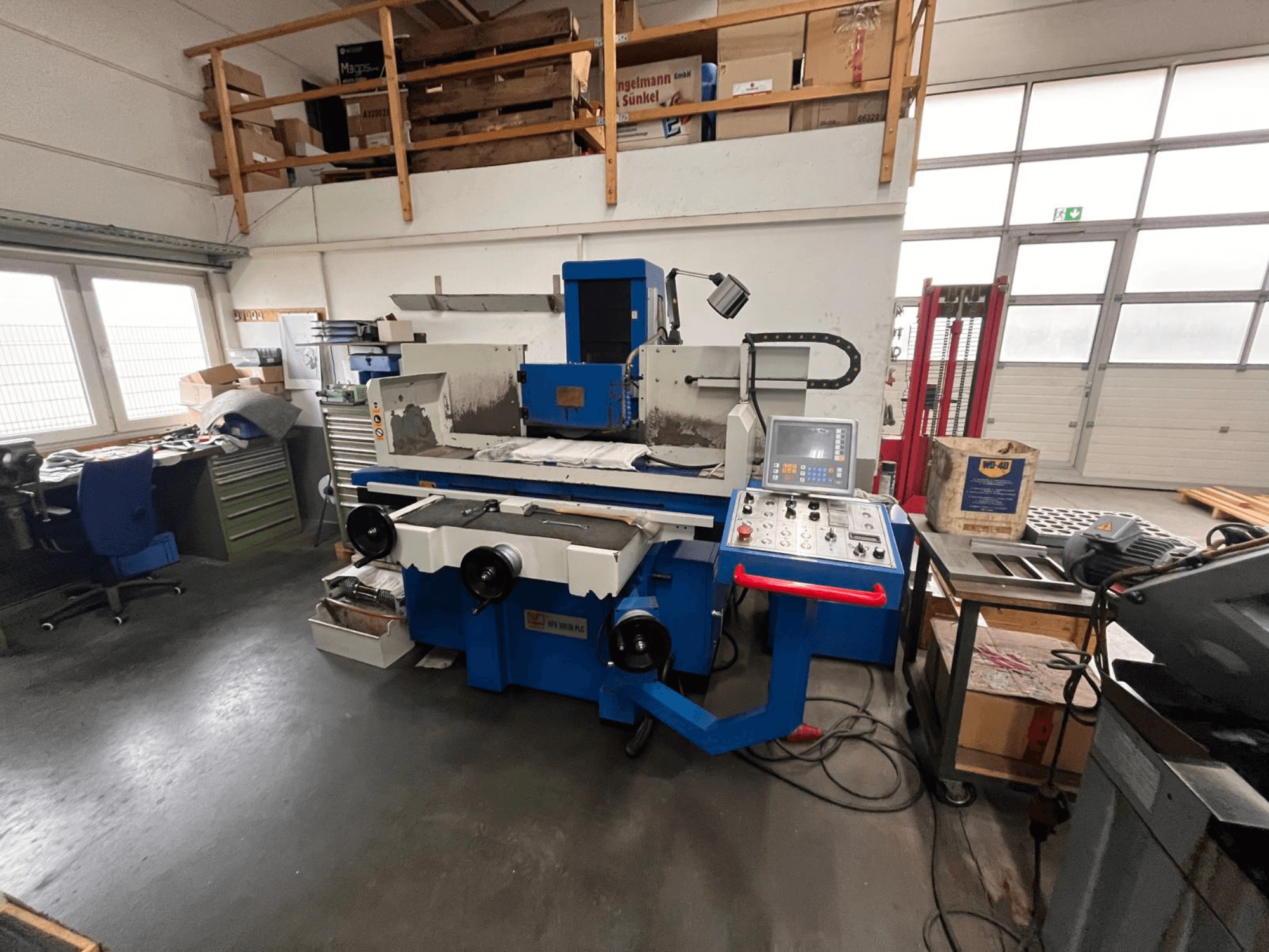 Zila virsmu slīpmašīna ar digitālo vadības paneli, skats no priekšpuses, kurā redzams darba vietas juceklis un zaļš instrumentu skapis.